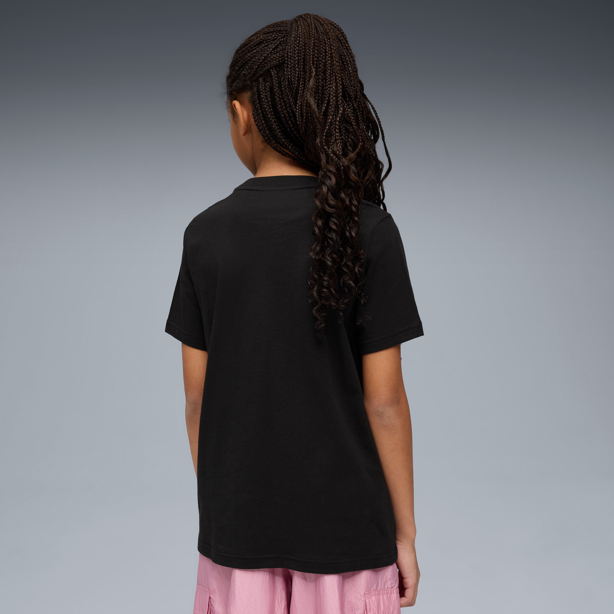 Modella di spalle con trecce, indossa t-shirt nera e pantaloncini rosa, su sfondo grigio.