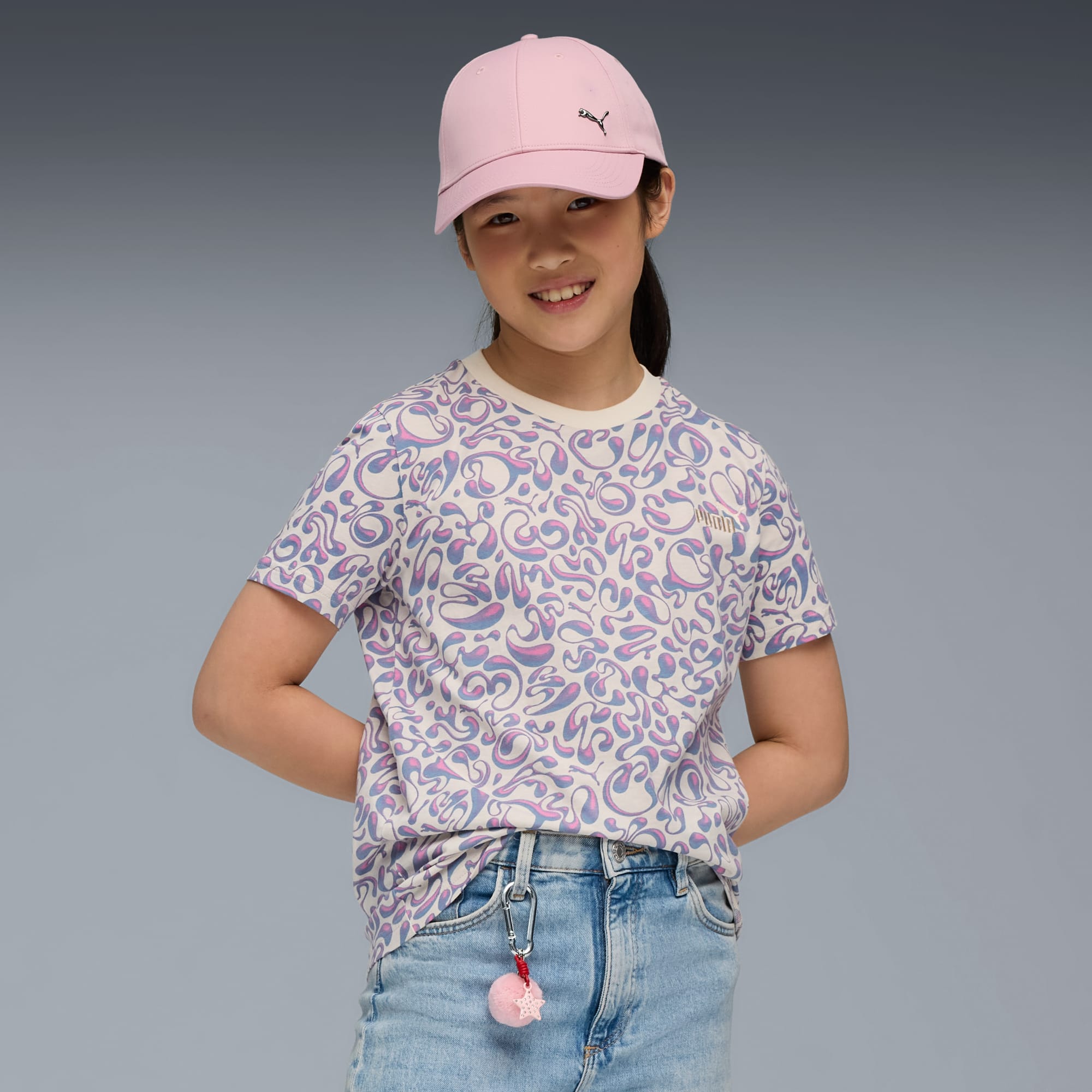 Una modella sorridente indossa una t-shirt fantasia, jeans e cappellino rosa su sfondo grigio.