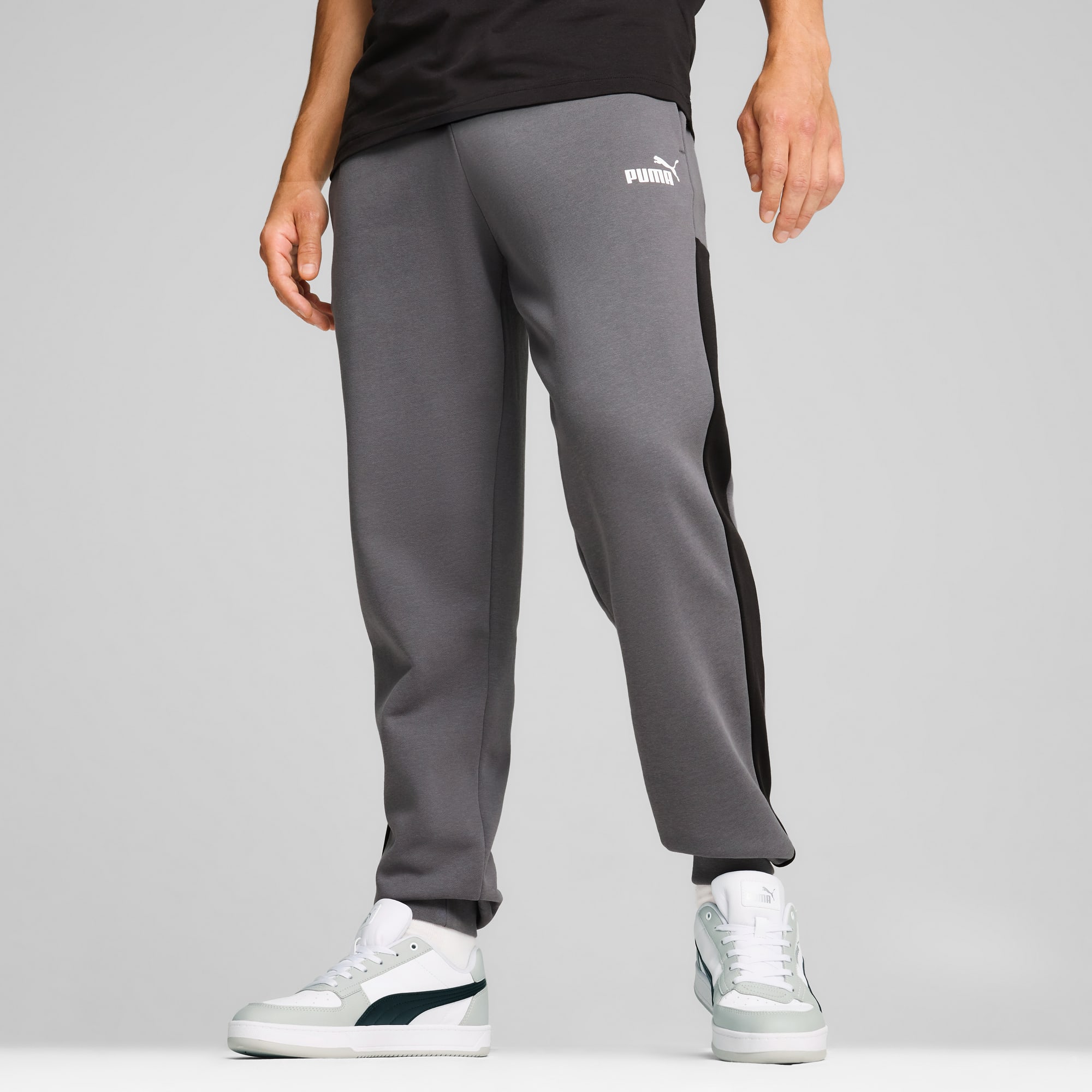 Een model staat tegen een grijze achtergrond, gekleed in een grijze broek en witte sneakers.