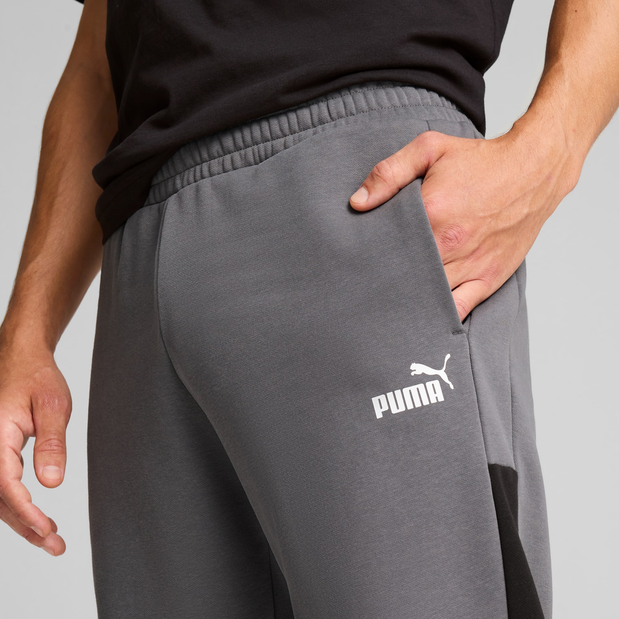 Een persoon met een hand in de zak draagt een grijze Puma broek tegen een grijze achtergrond.