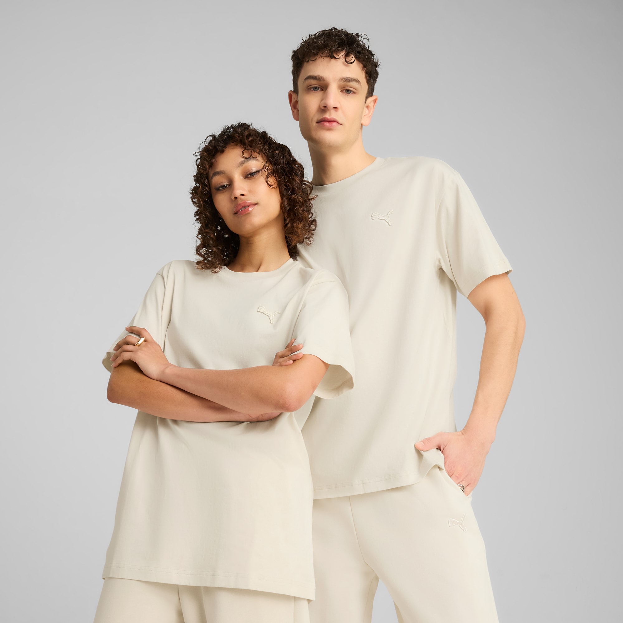 Twee modellen poseren tegen een grijze achtergrond, gekleed in bijpassende, neutraal getinte t-shirts en broeken.