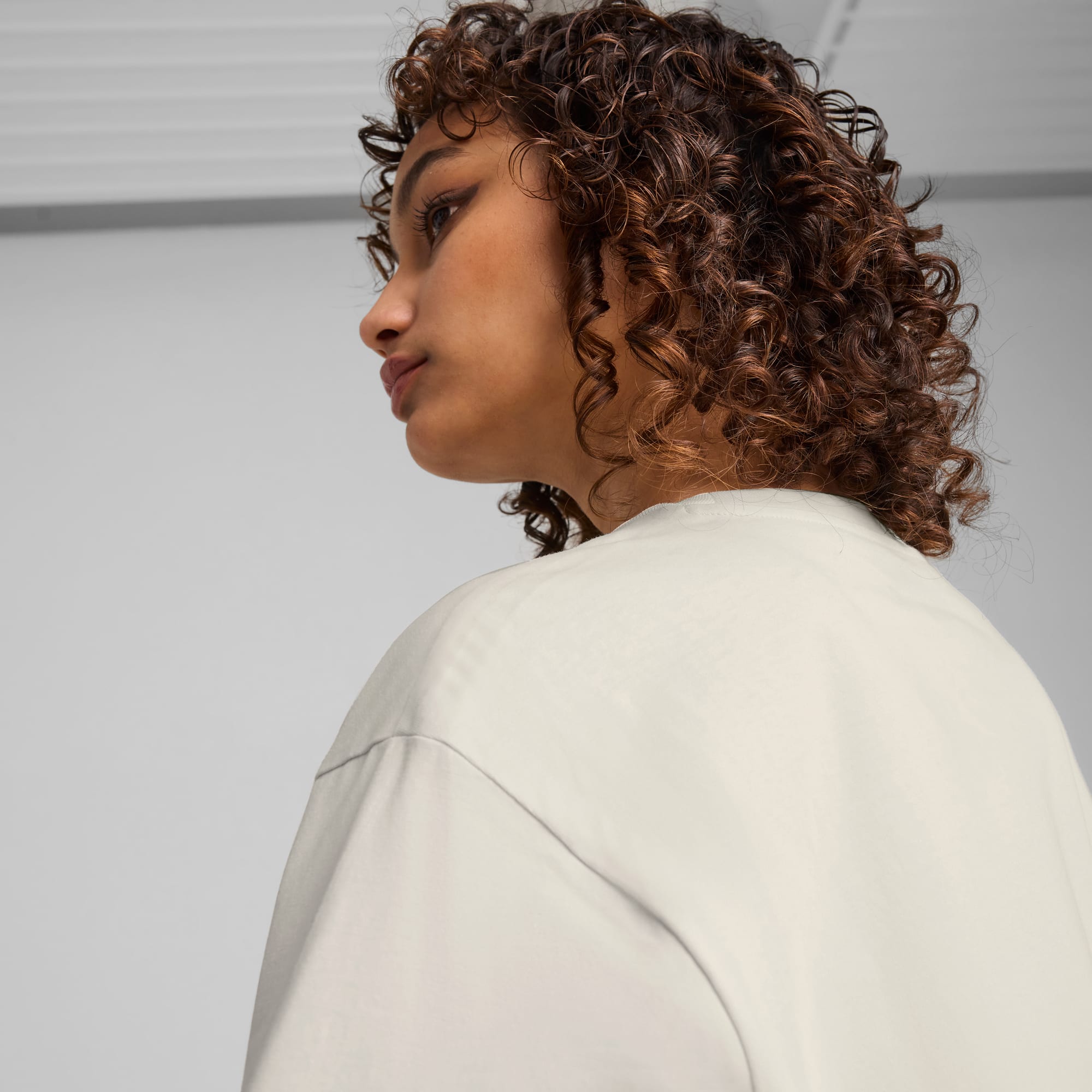 Against a grey backdrop, a model with curly hair wears a light-coloured t-shirt.