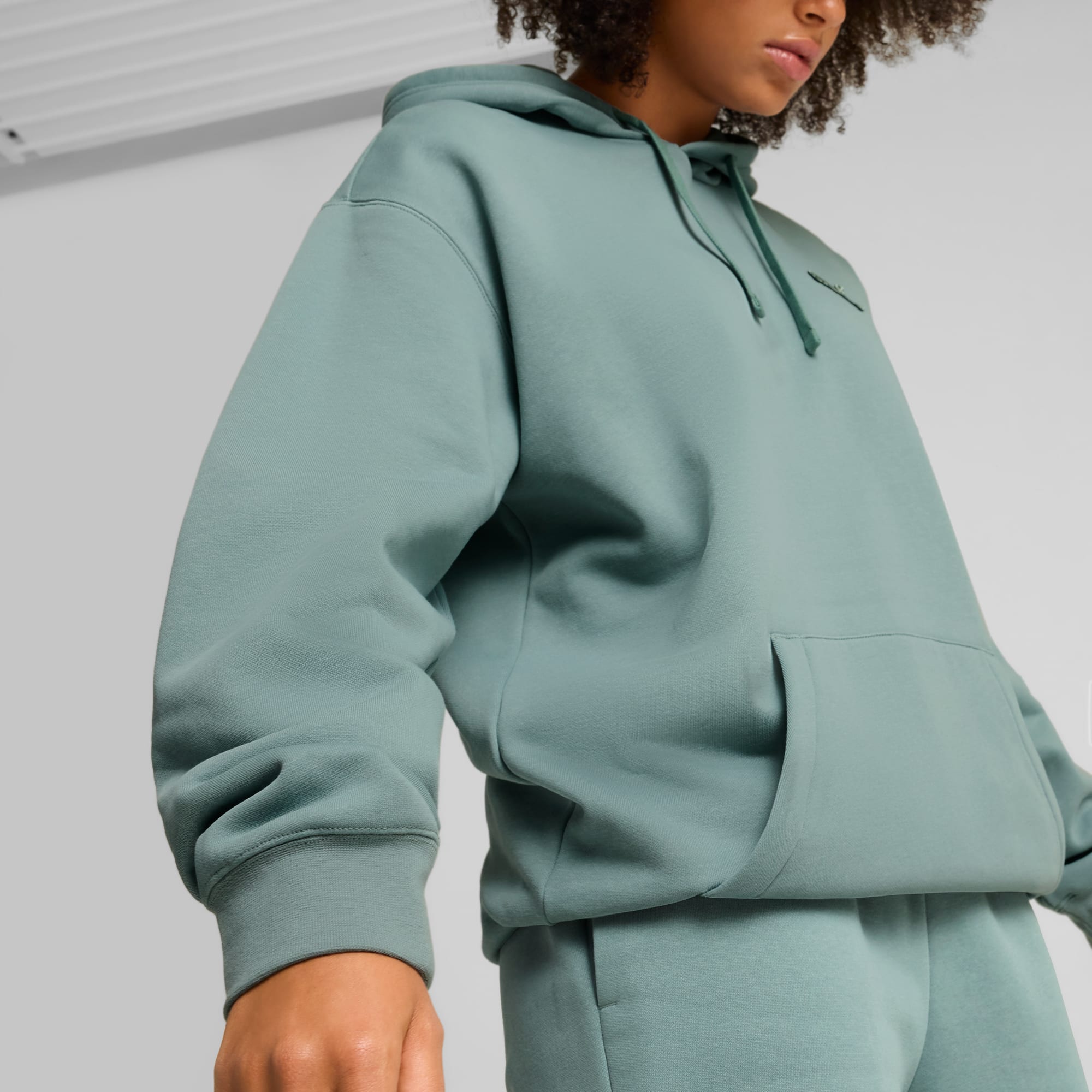 Model draagt een blauwgroene hoodie en bijpassende joggingbroek tegen een neutrale achtergrond.