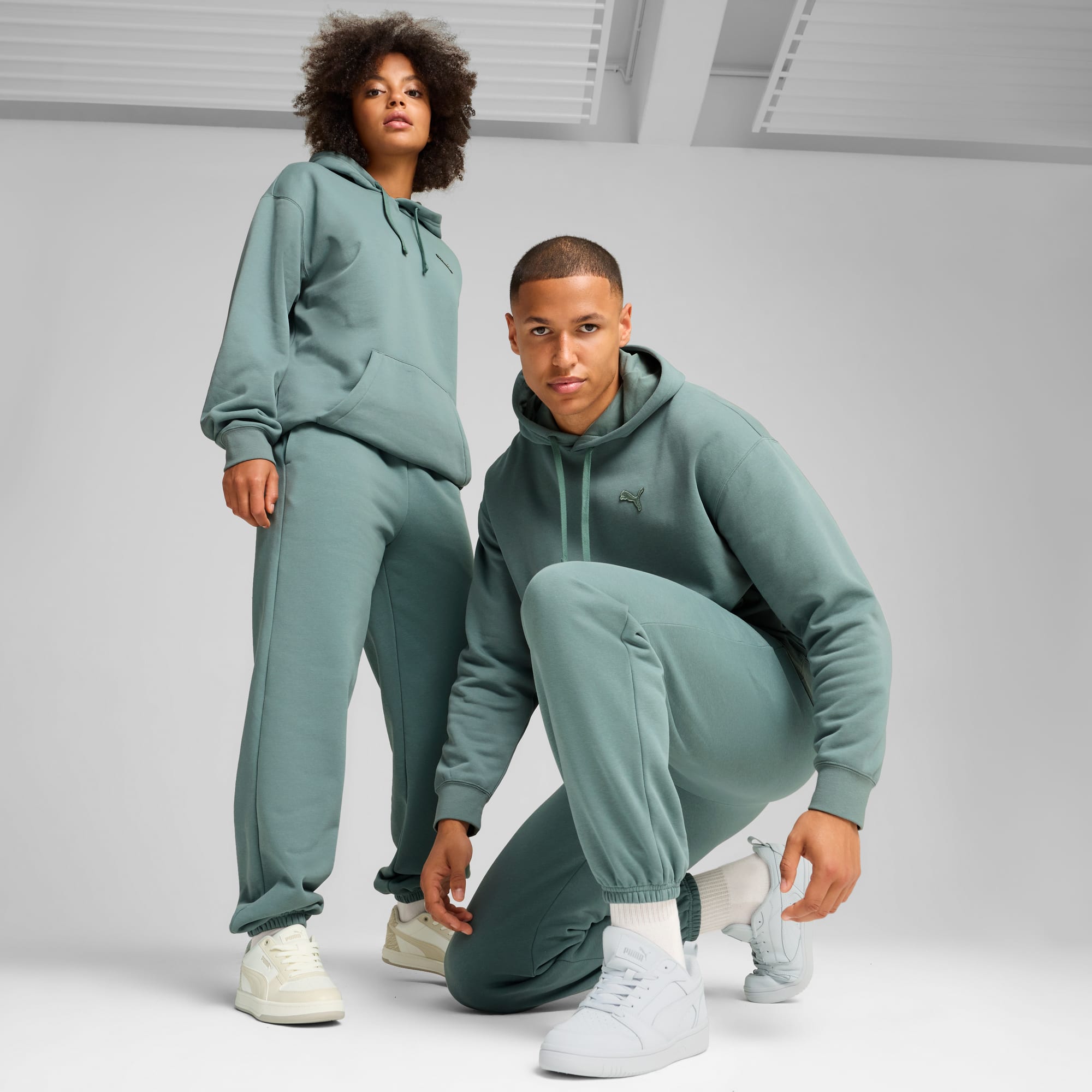 Zwei Models posieren vor grauem Hintergrund in passenden, petrolfarbenen Hoodies und Jogginghosen.