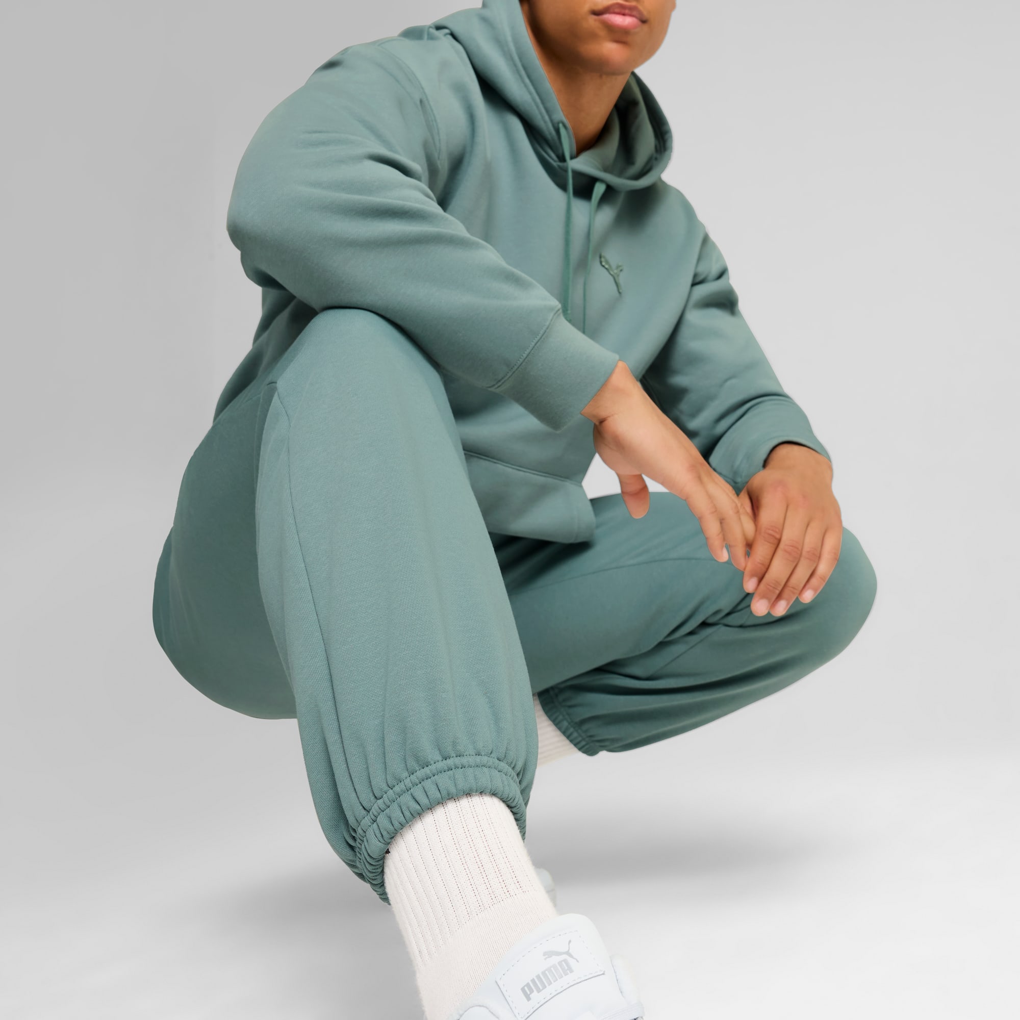 Vor grauem Hintergrund kauert ein Model in grünem Hoodie und passender Jogginghose.