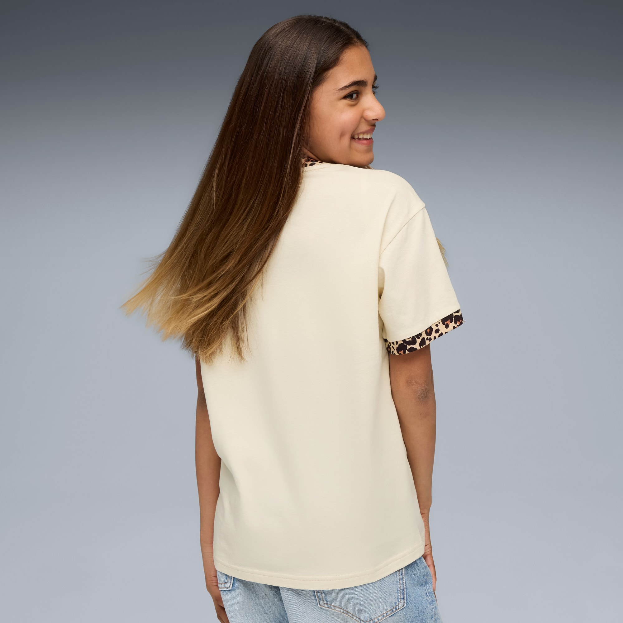 Ein Model mit langen Haaren lächelt und trägt ein cremefarbenes T-Shirt mit Animal-Print vor Grau.