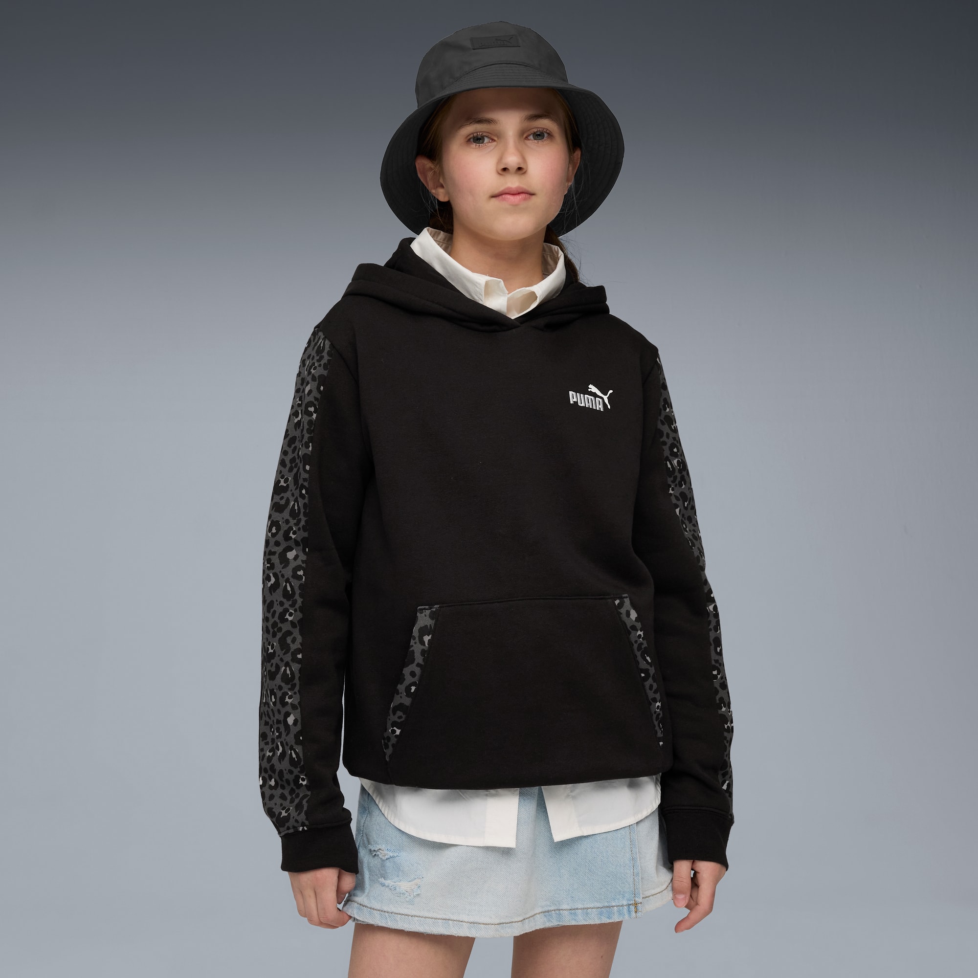 Sur fond gris, un mannequin porte un chapeau, un sweat à capuche noir avec un logo et une jupe en jean.