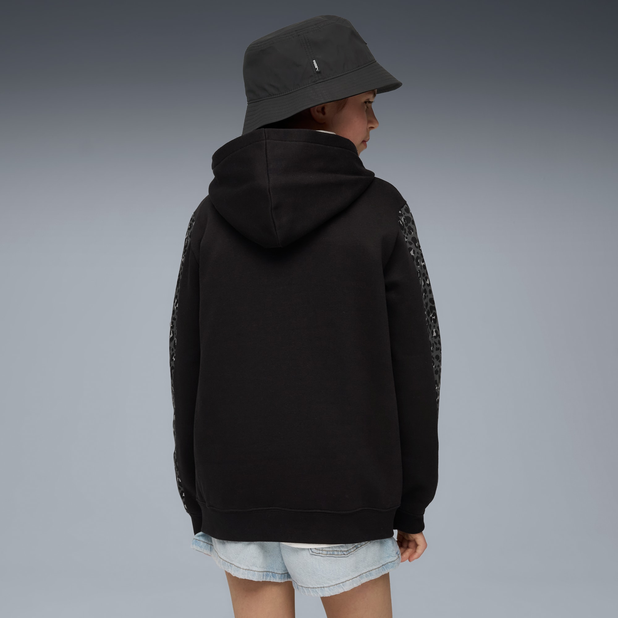 Sur fond gris, un mannequin porte un sweat à capuche noir, un short en jean et un chapeau, vu de dos.