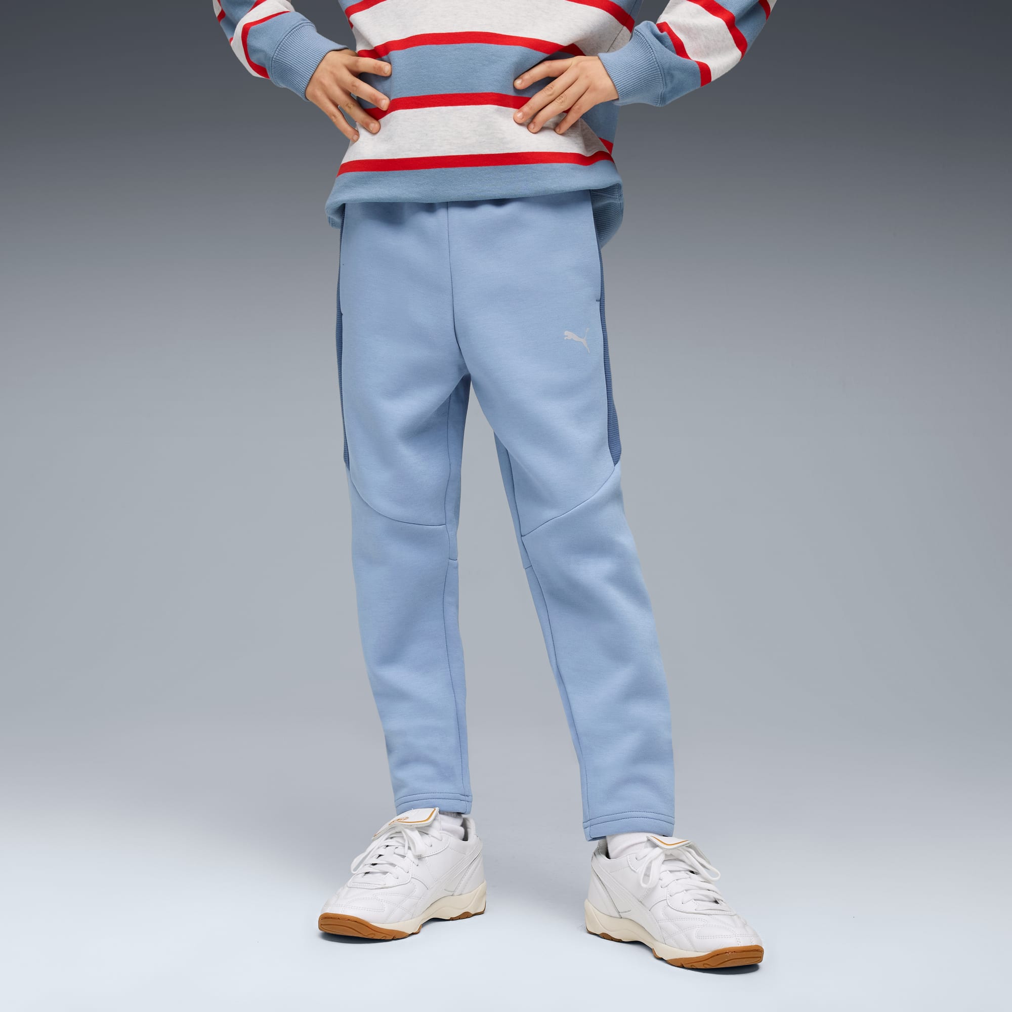 Een model poseert tegen een grijze achtergrond, gekleed in een lichtblauwe broek en witte sneakers.