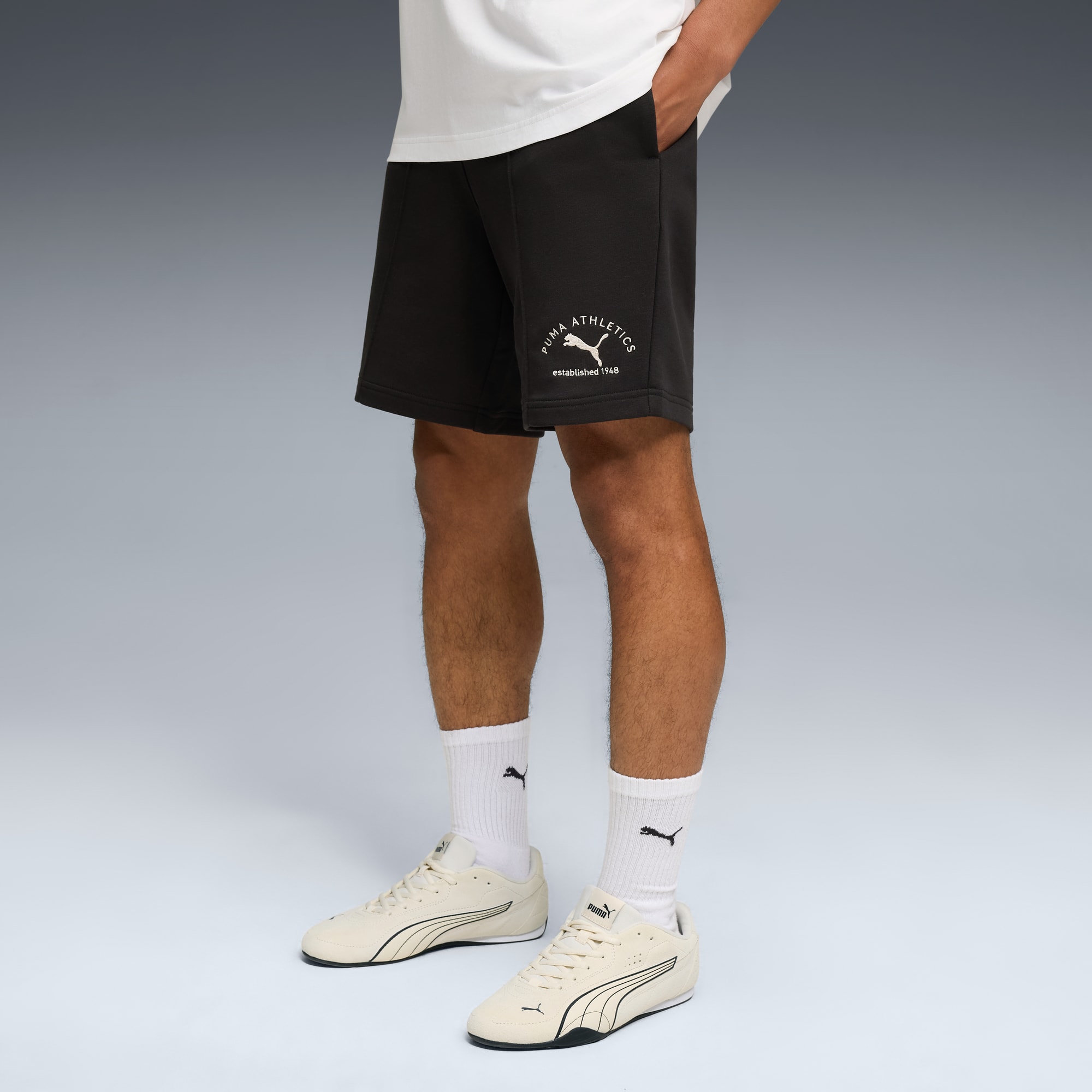 Sur fond gris, un modèle porte un t-shirt blanc, un short foncé, des chaussettes blanches et des baskets claires.
