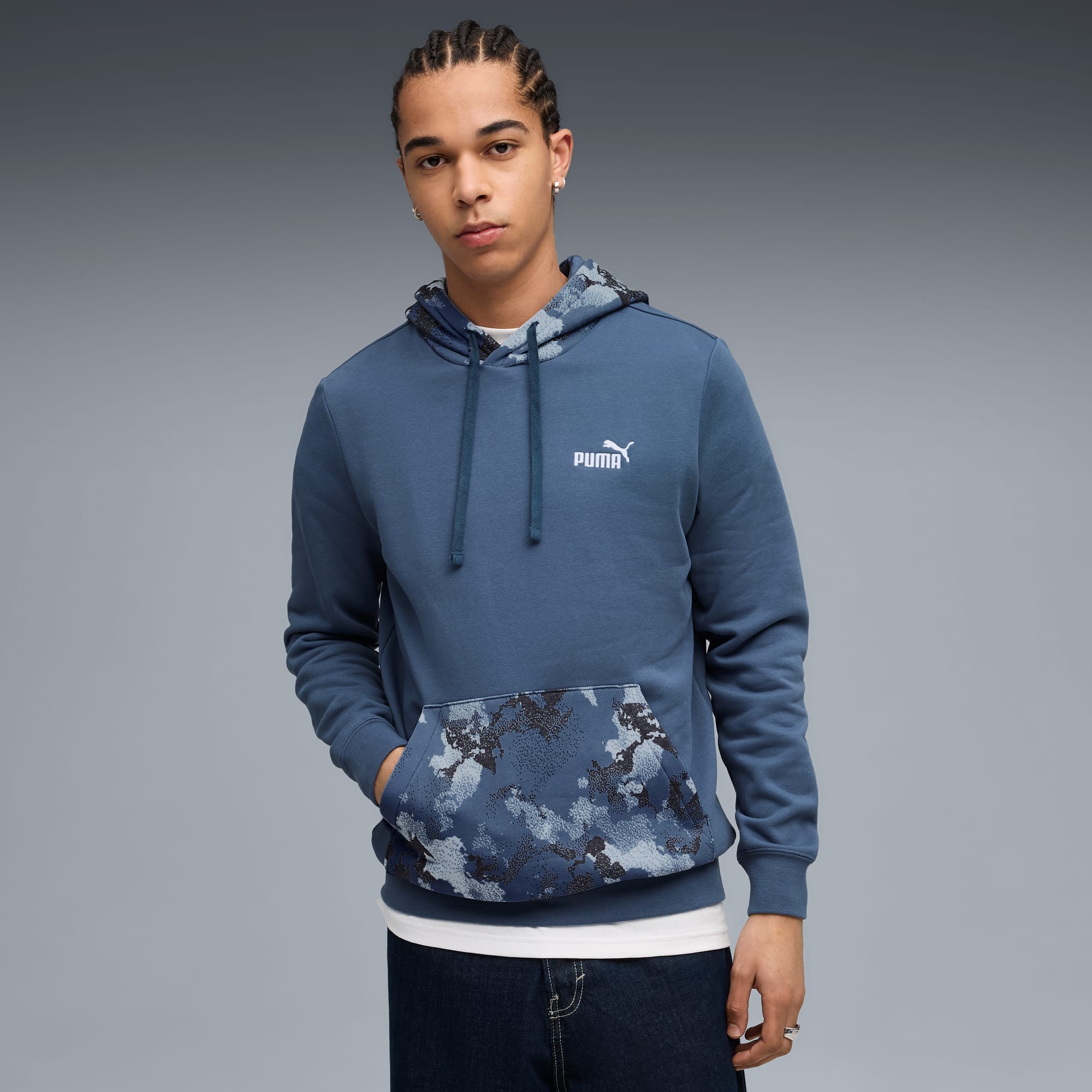 Sur fond gris, un modèle pose en sweat à capuche Puma bleu avec motifs sur la capuche et la poche.