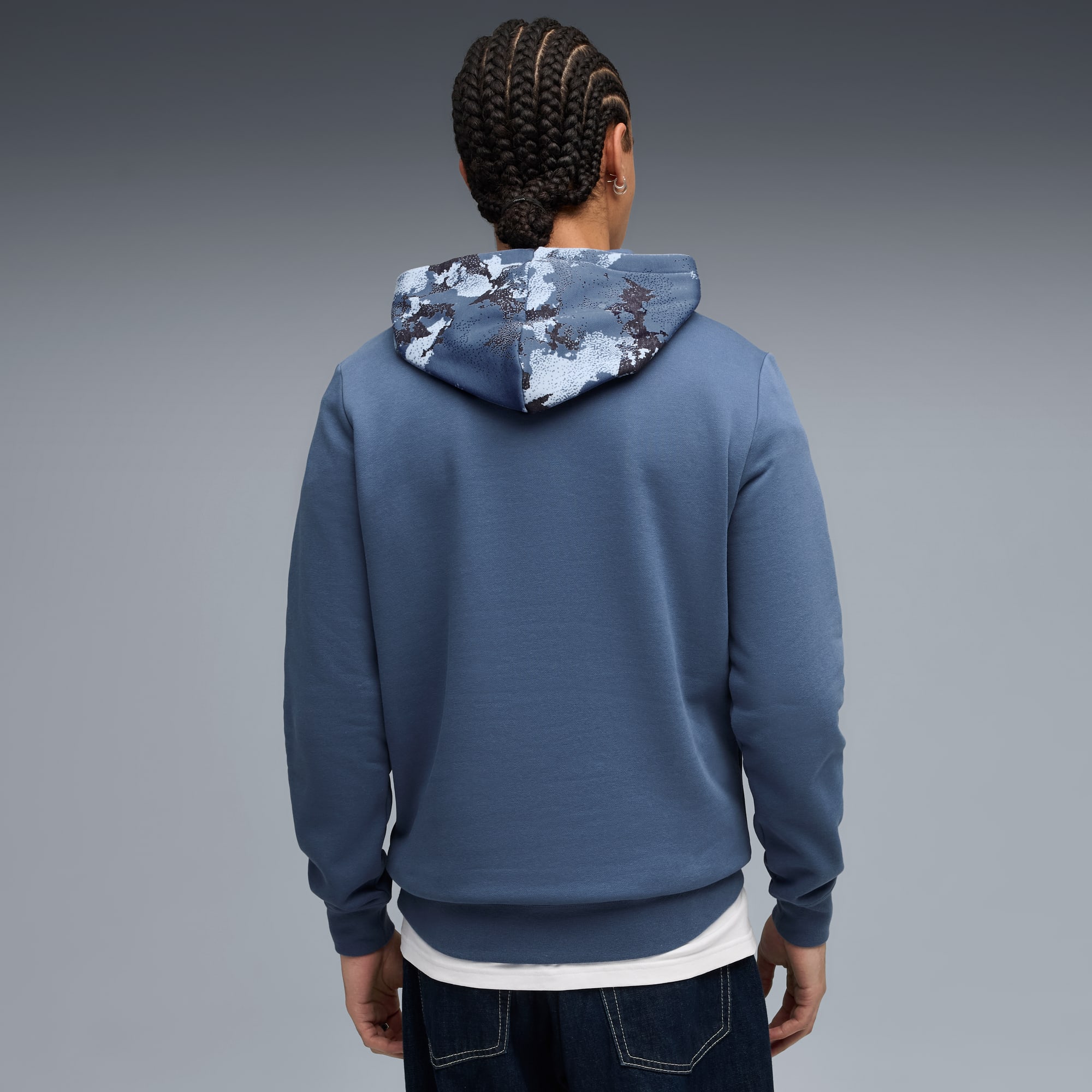 Un modèle, de dos sur fond gris, porte un sweat à capuche bleu avec une capuche à motifs et un jean.