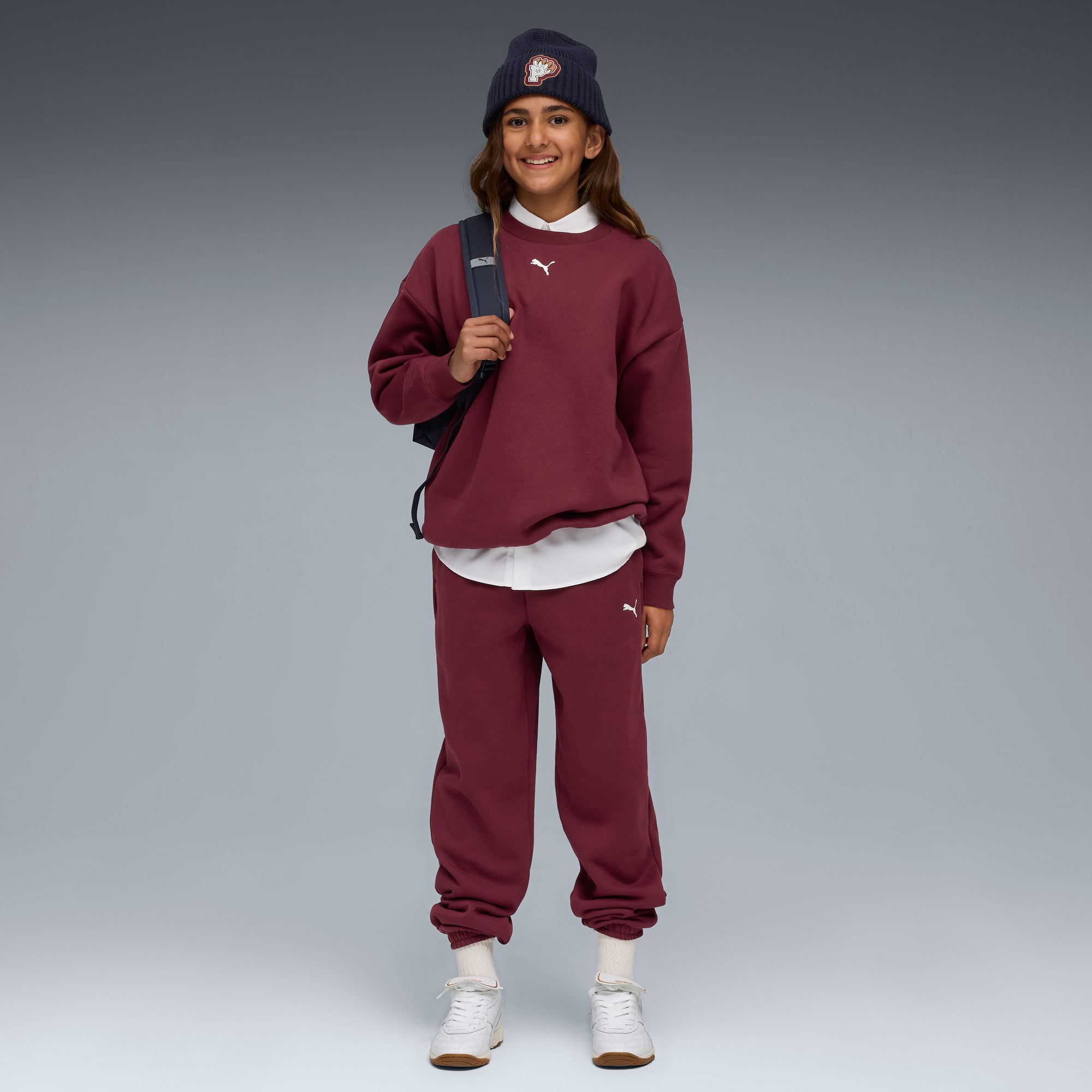 Tegen een grijze achtergrond draagt een model een muts, bordeaux trainingspak en sneakers.