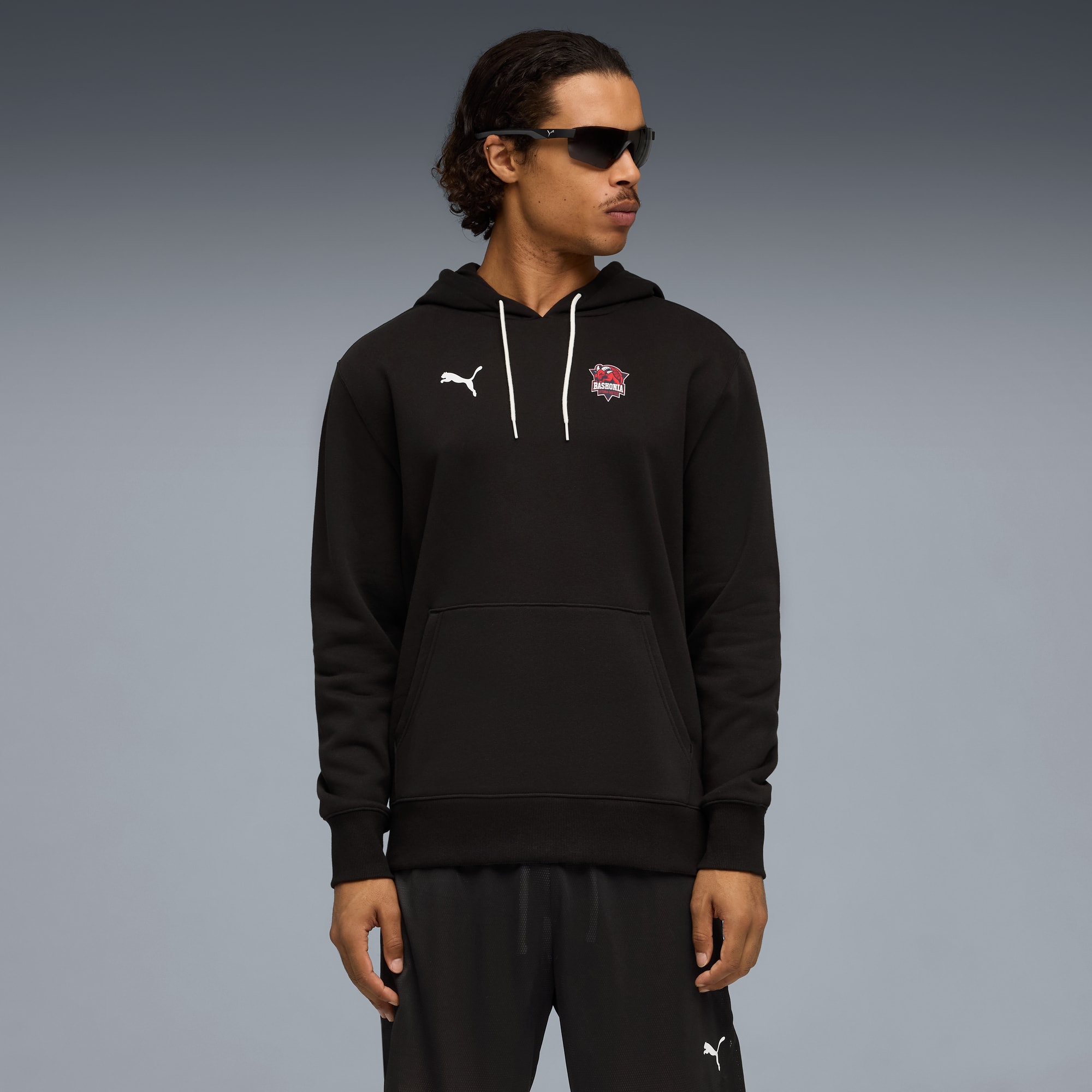 Sur fond gris, un mannequin pose avec des lunettes de soleil, un sweat à capuche noir et un bas assorti.