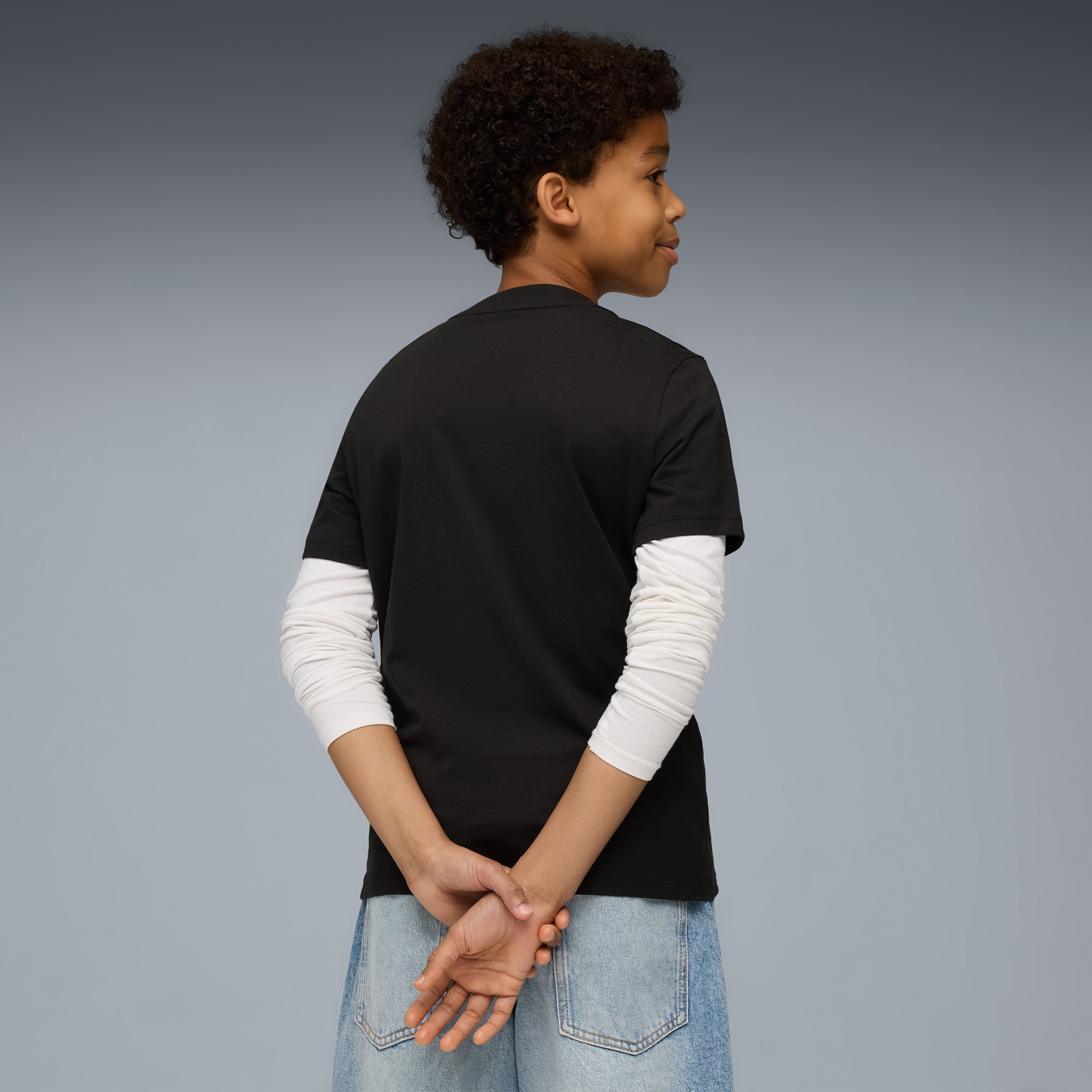 Un enfant aux cheveux bouclés, sur fond gris, porte un t-shirt noir, un haut à manches longues et un short en jean.