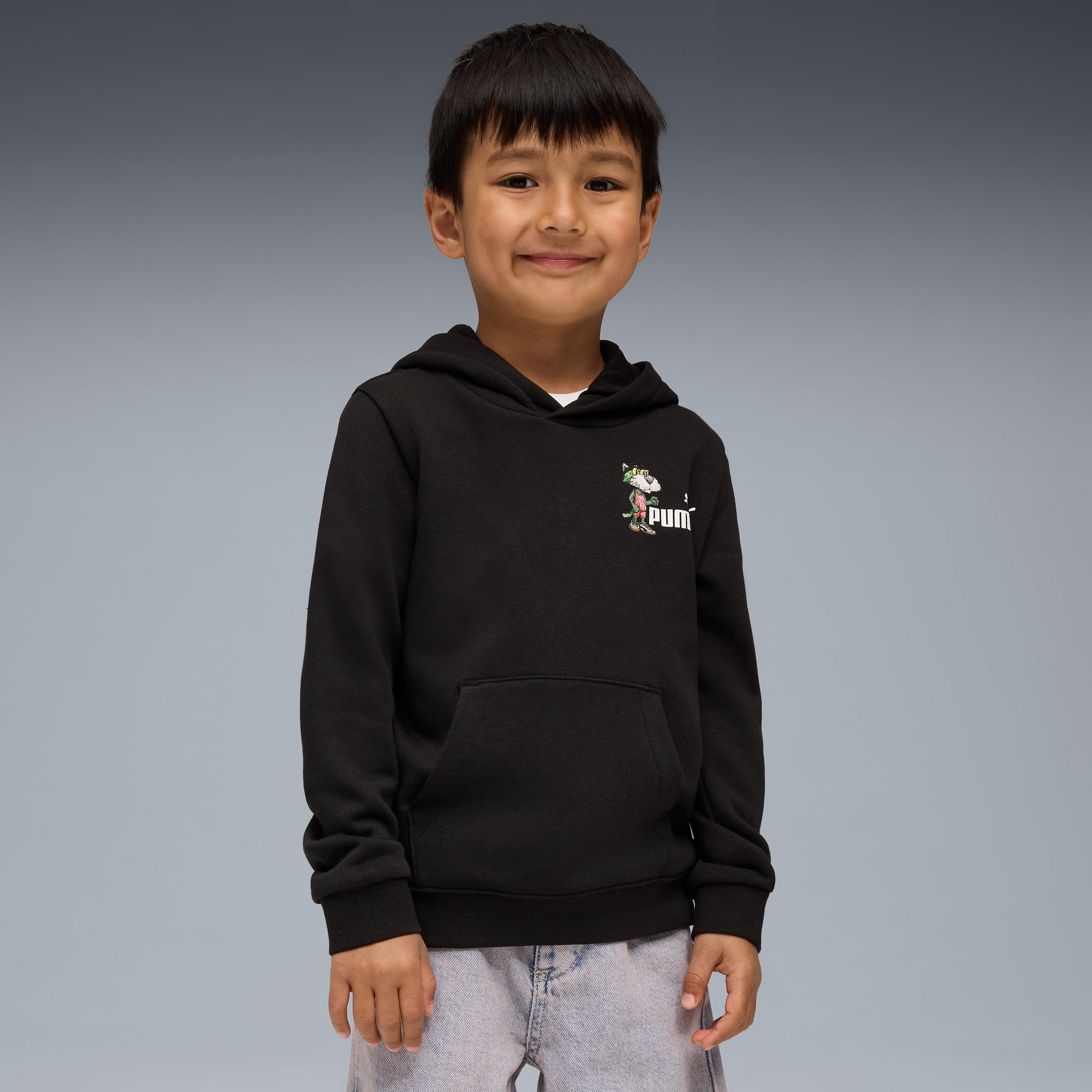 Un enfant souriant pose sur fond gris, portant un sweat à capuche noir et un jean clair.