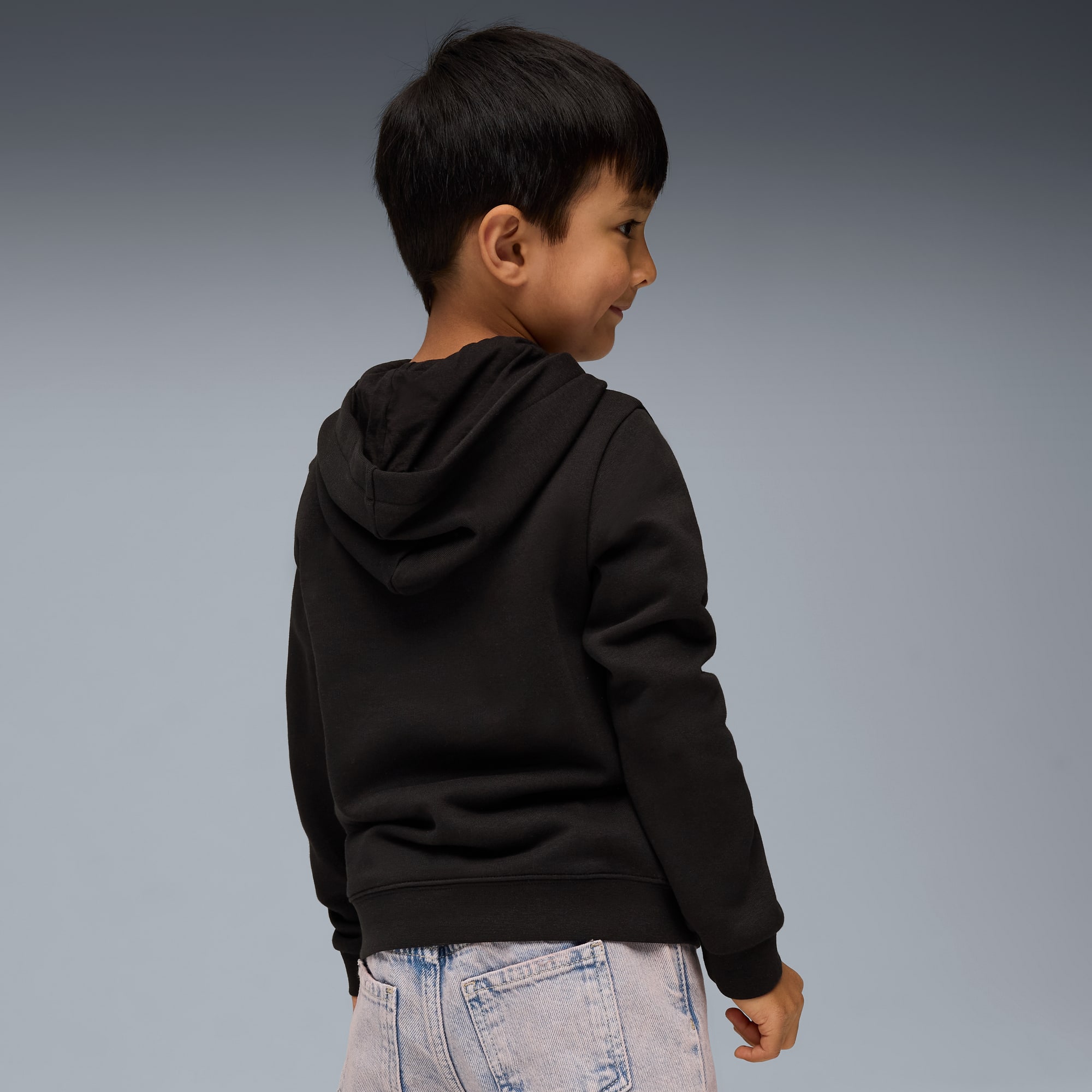 Un enfant en sweat à capuche noir et jean regarde sur le côté sur fond gris.