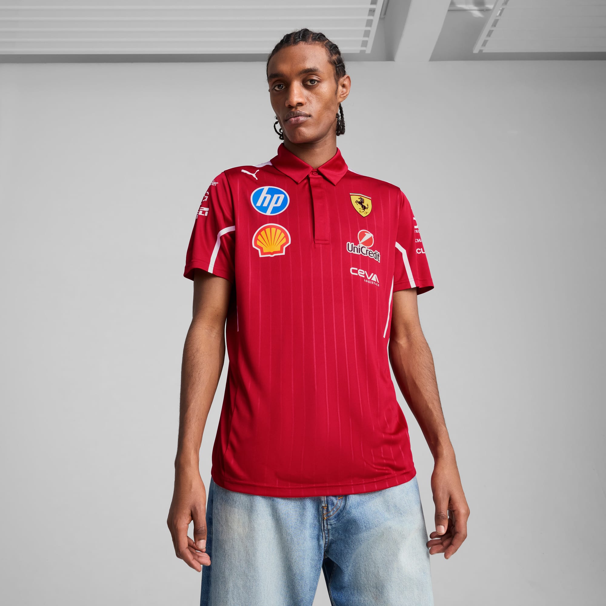 Tegen een grijze achtergrond poseert een model in een rood poloshirt met logo's en lichte jeans.