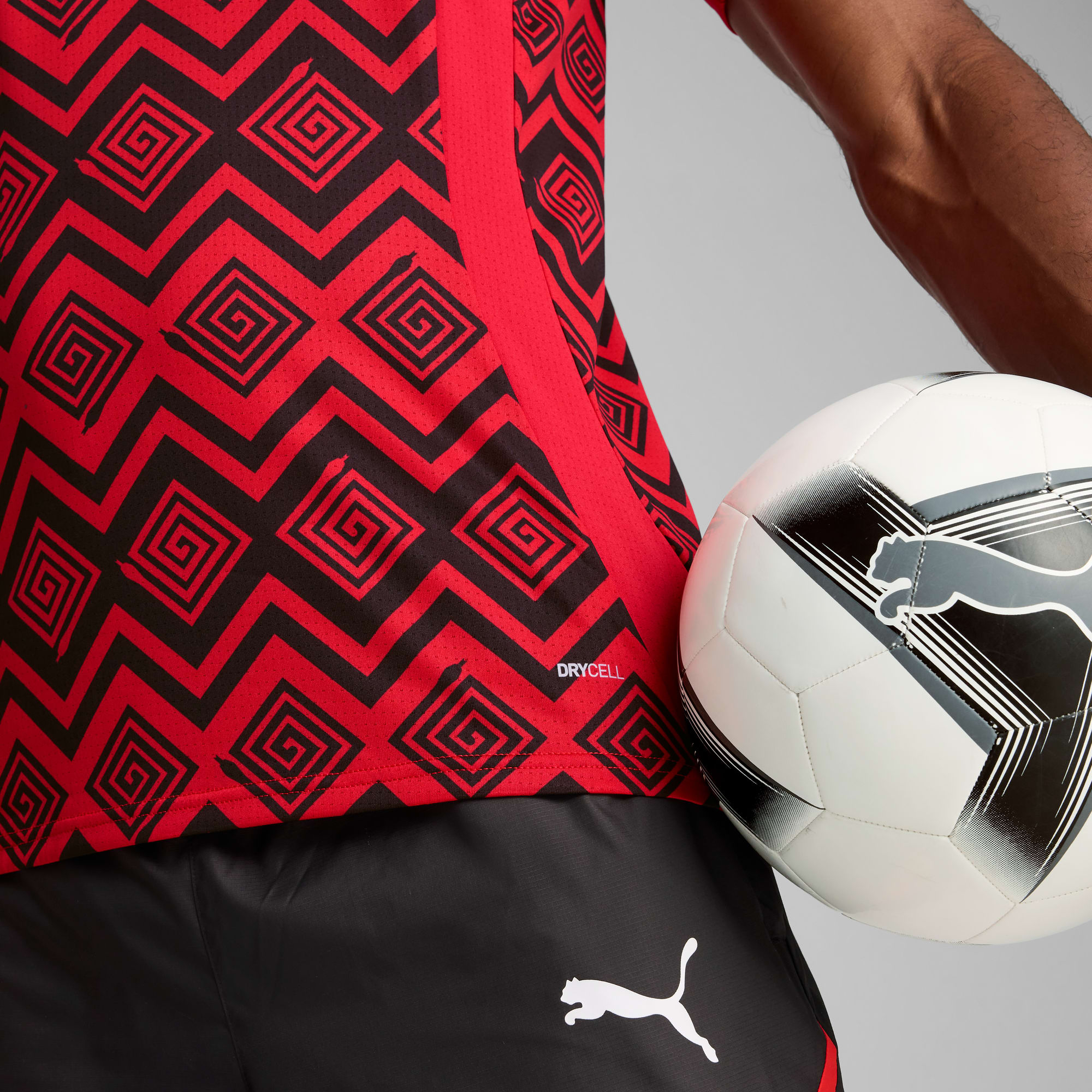 Een model houdt een voetbal vast tegen een grijze achtergrond, gekleed in een rood shirt met patroon en zwarte shorts.