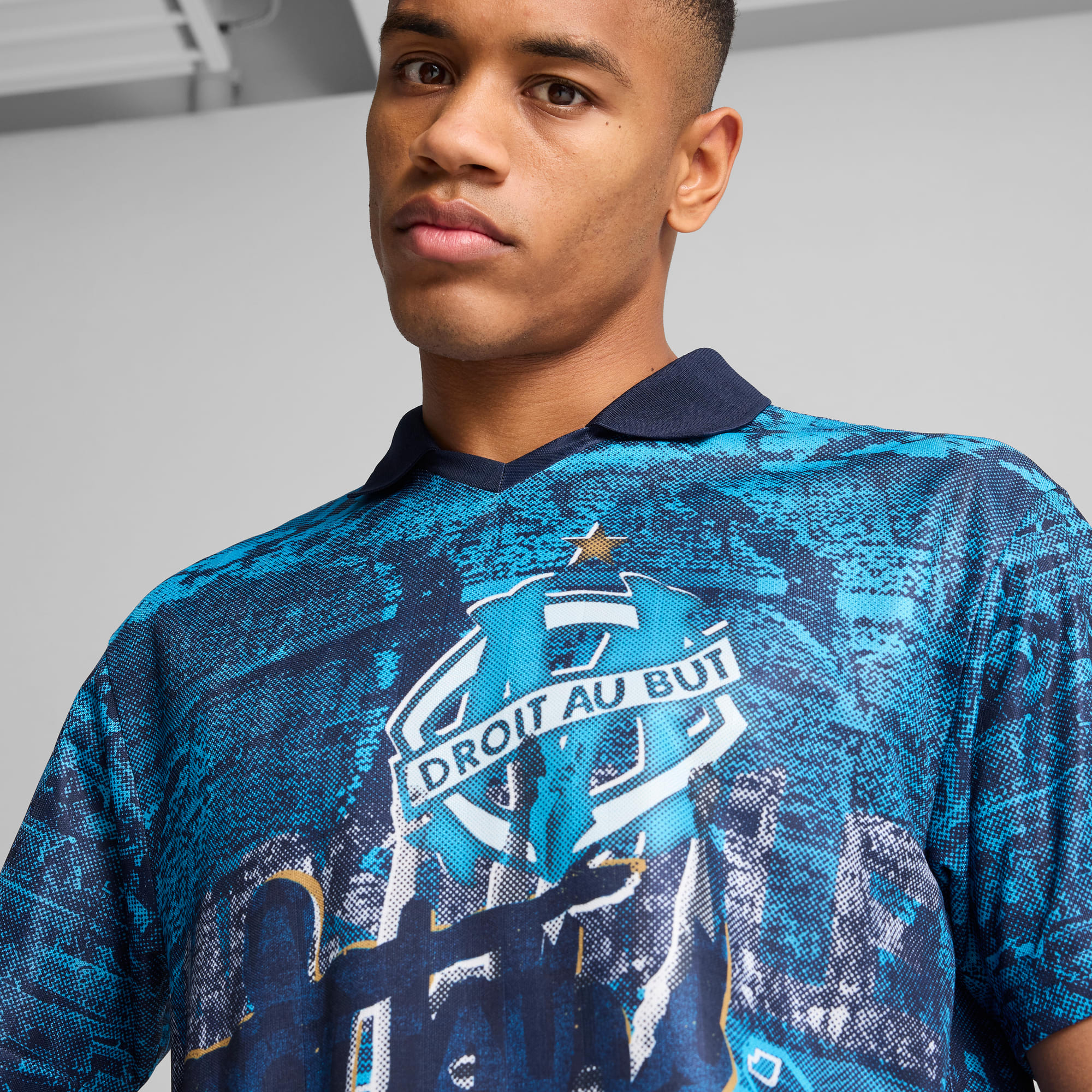 Vor grauem Hintergrund trägt ein Model ein blau gemustertes Fussballtrikot mit marineblauem Kragen und Logo.