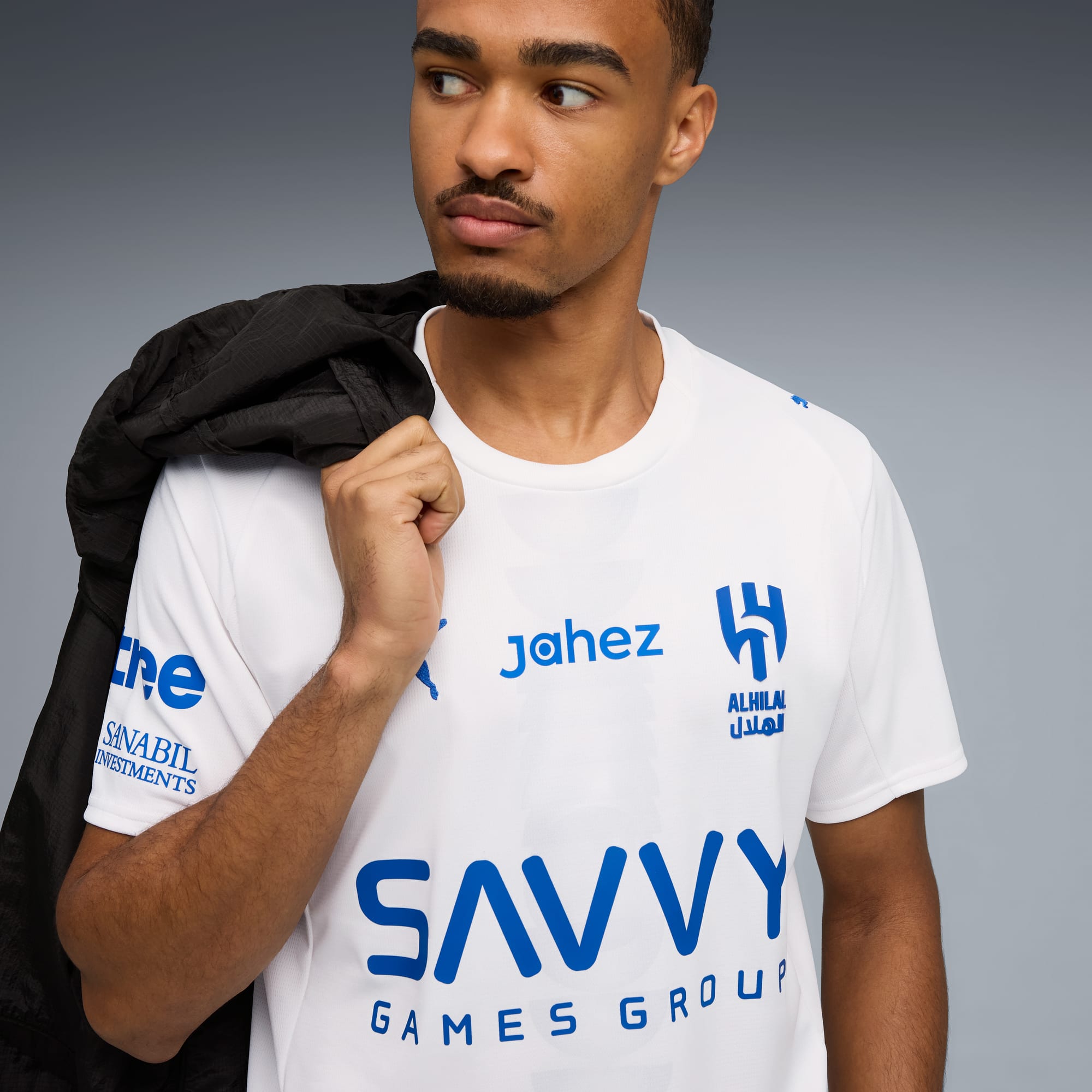 Sur fond gris, un mannequin porte un t-shirt blanc avec des logos bleus et tient une veste noire.