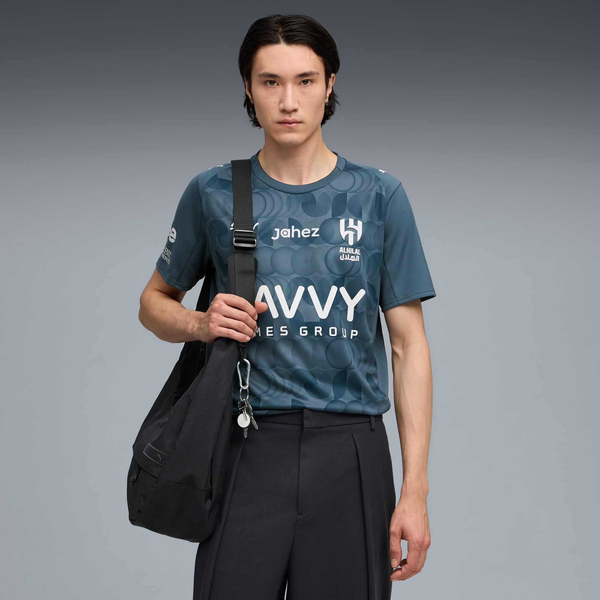 Su sfondo grigio, un modello posa con una t-shirt fantasia, pantaloni scuri e borsa a tracolla.