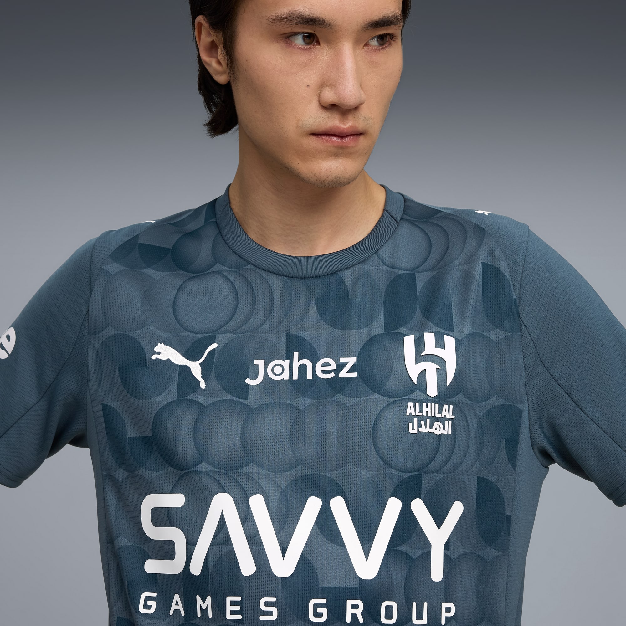 Un modello indossa una t-shirt grigia a motivi con loghi di sponsor su sfondo grigio.