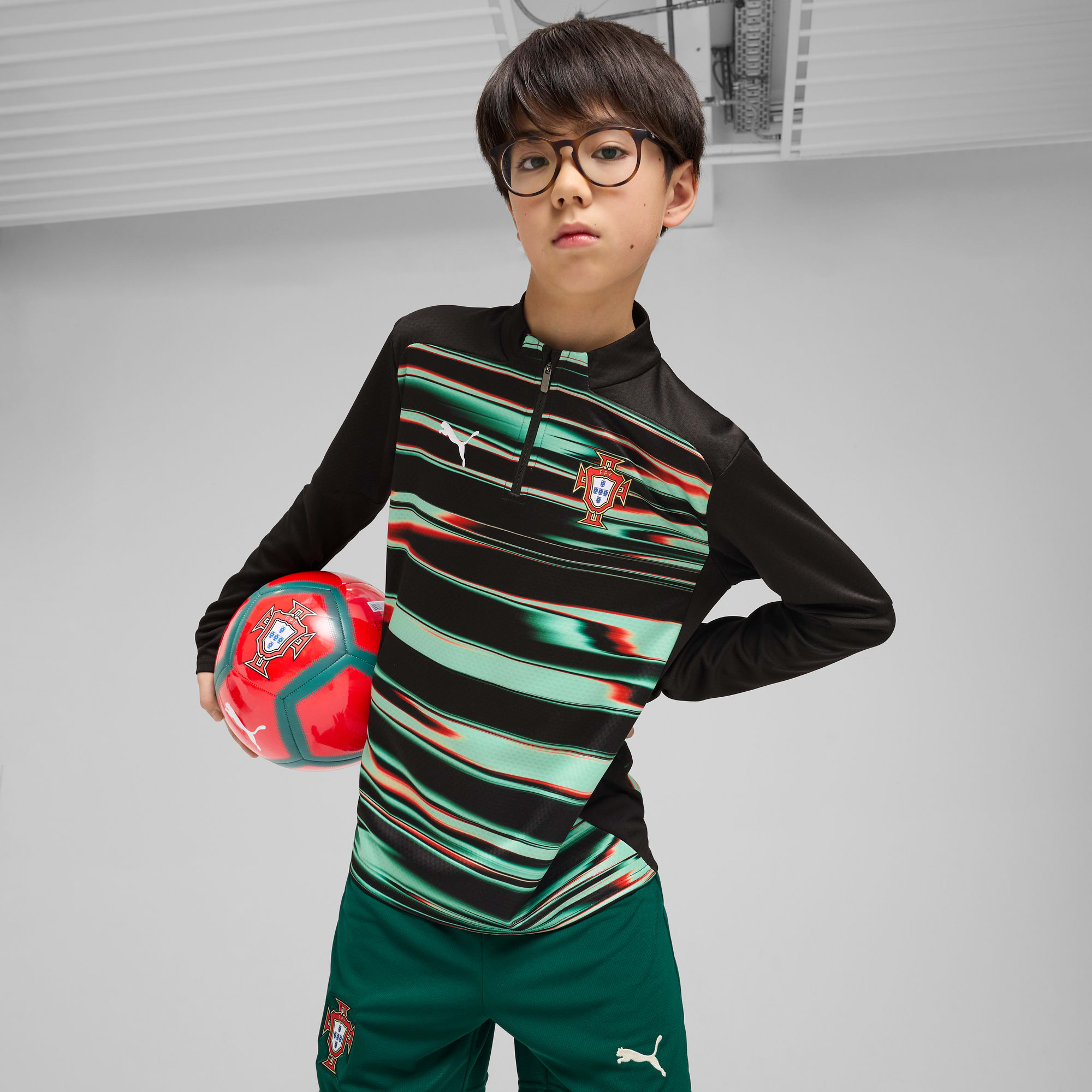 Ein Junge mit Brille posiert mit einem Fußball vor grauem Hintergrund in Sportbekleidung.
