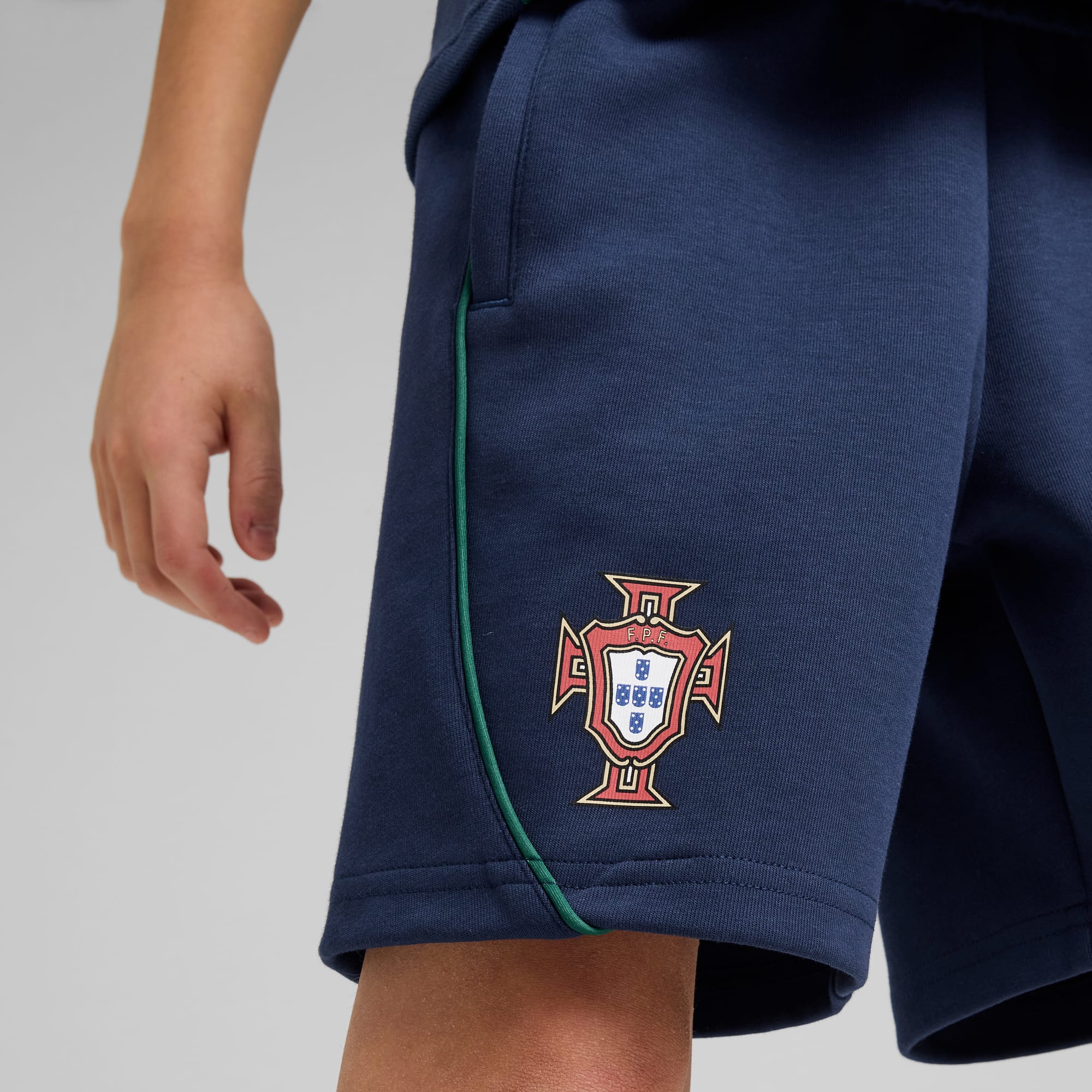 Against grey, a person wears navy shorts with the Portuguese emblem and green trim.