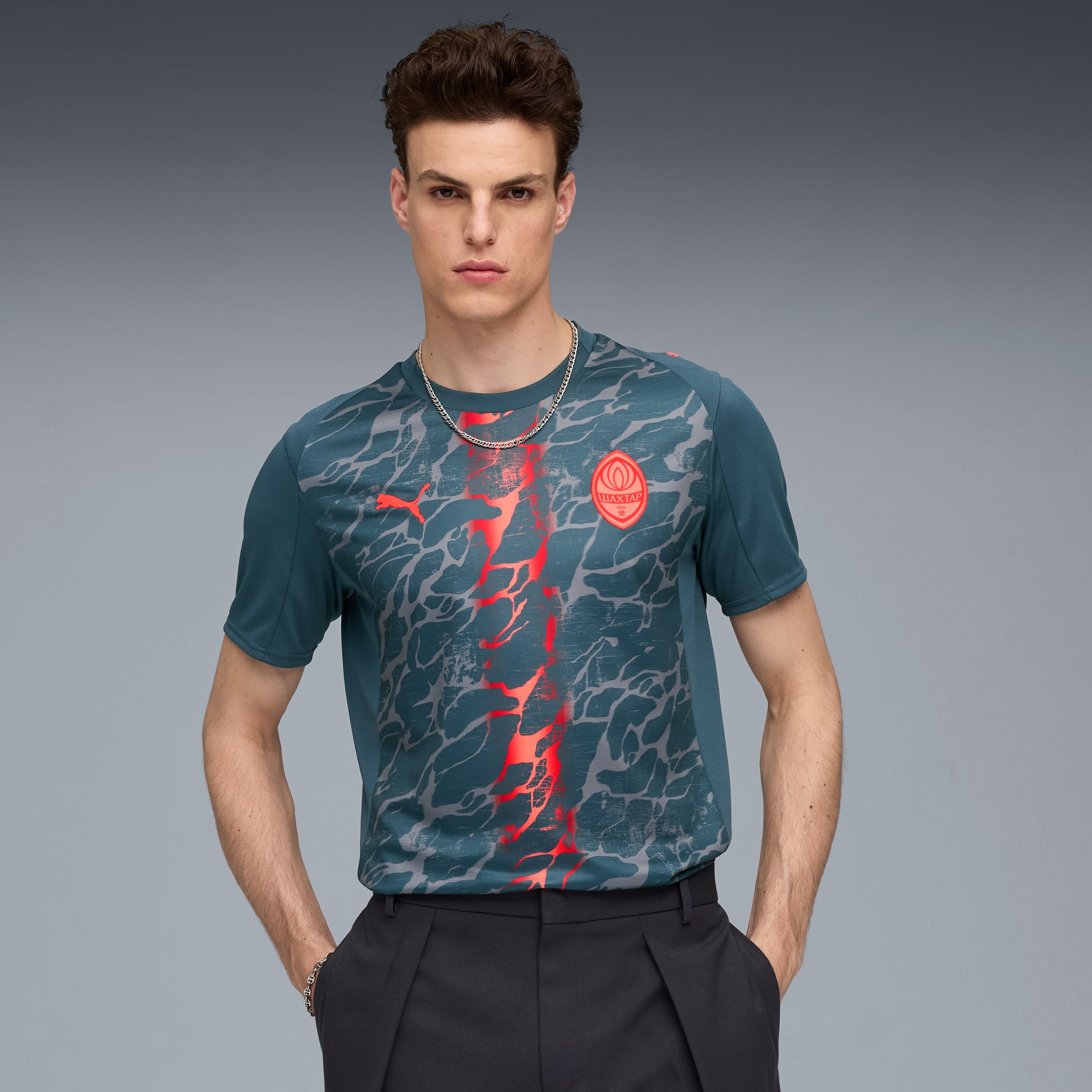 Een model poseert tegen grijs, gekleed in een blauwgroen T-shirt met patroon en een donkere broek.