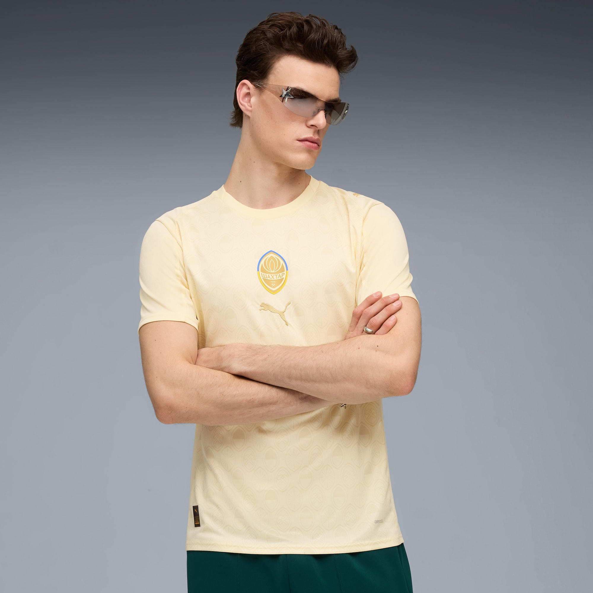 Un modello con occhiali da sole posa con una t-shirt gialla su sfondo grigio.
