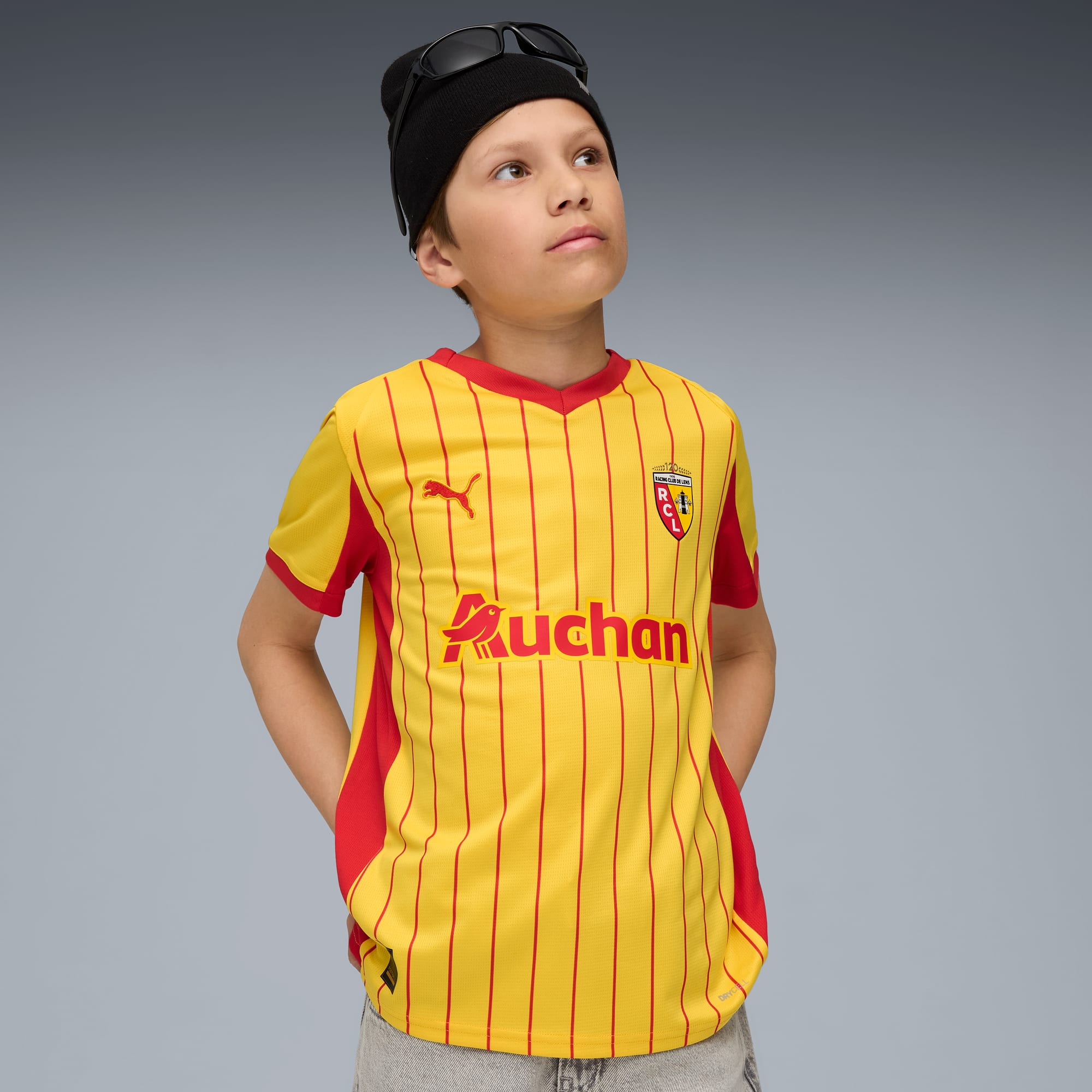 Un enfant pose sur fond gris, portant un maillot jaune et rouge, un bonnet noir et des lunettes.