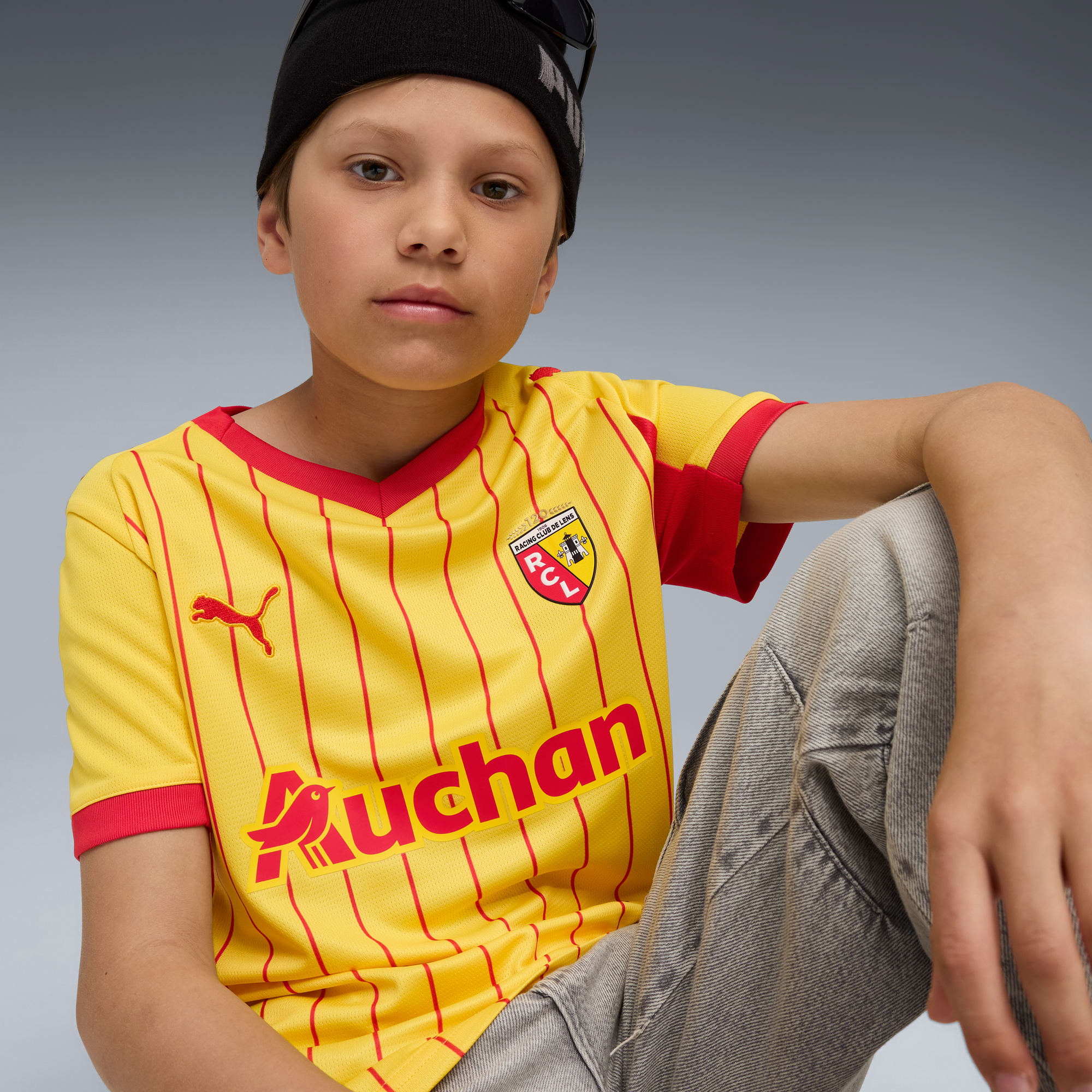 Un modèle est assis sur un fond gris, portant un maillot jaune et rouge avec un bonnet noir.