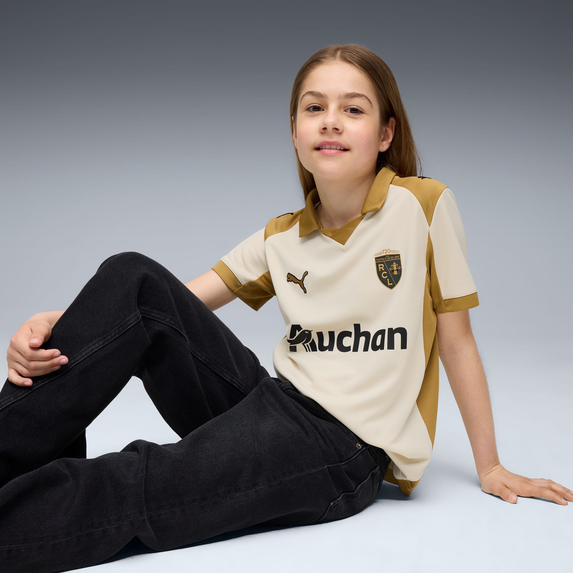 Un modello è seduto su uno sfondo grigio, indossa una maglietta color crema e oro e jeans scuri.