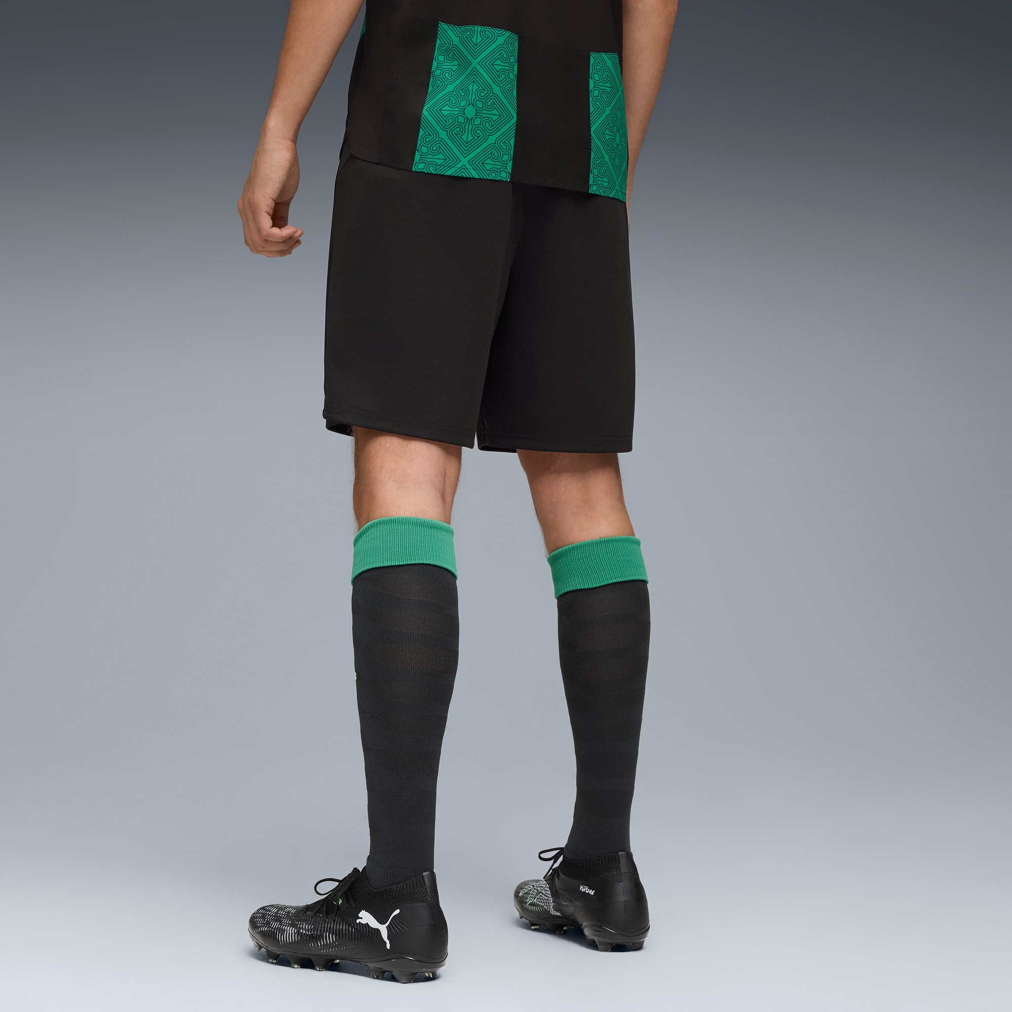 Sur fond gris, un modèle porte un short noir, un t-shirt, des chaussettes à bordure verte et des chaussures.