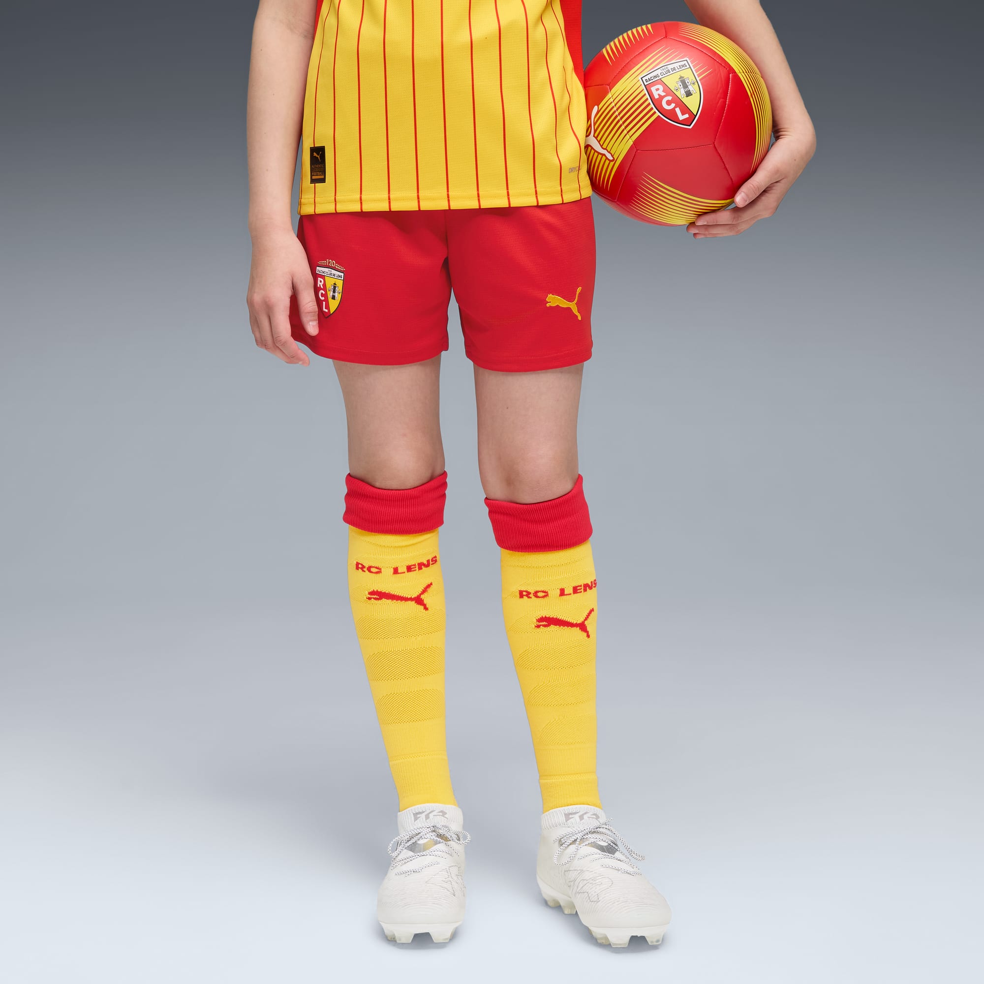 Su sfondo grigio, modello tiene un pallone indossando kit calcio giallo e rosso e scarpe da ginnastica.