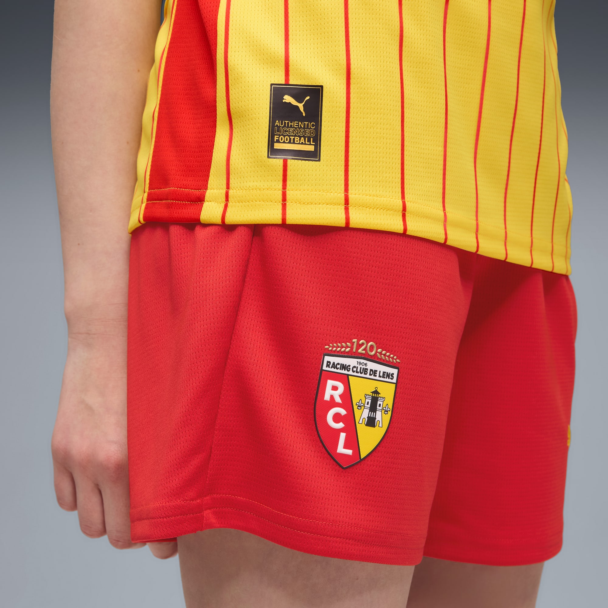 Su sfondo grigio, un modello indossa una maglia gialla e rossa e pantaloncini rossi con il logo del Racing Club de Lens.