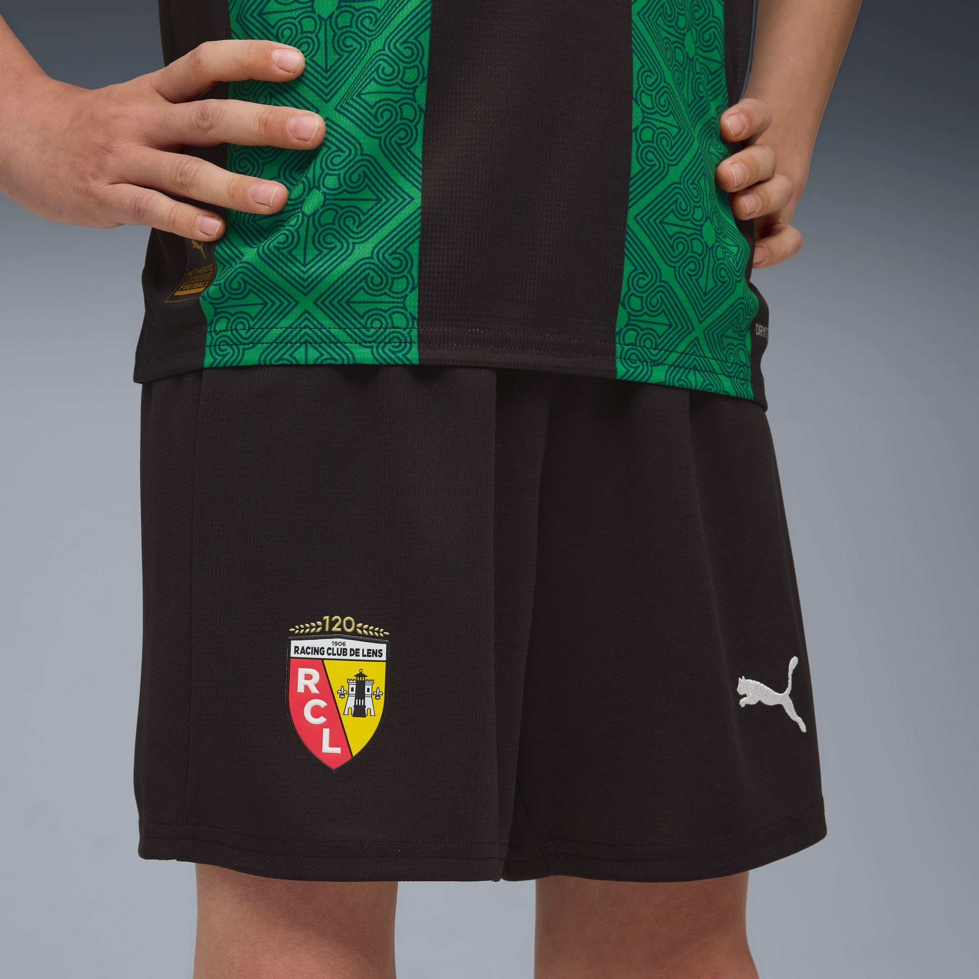 Un modello posa su uno sfondo grigio indossando una maglia verde e nera e pantaloncini neri.