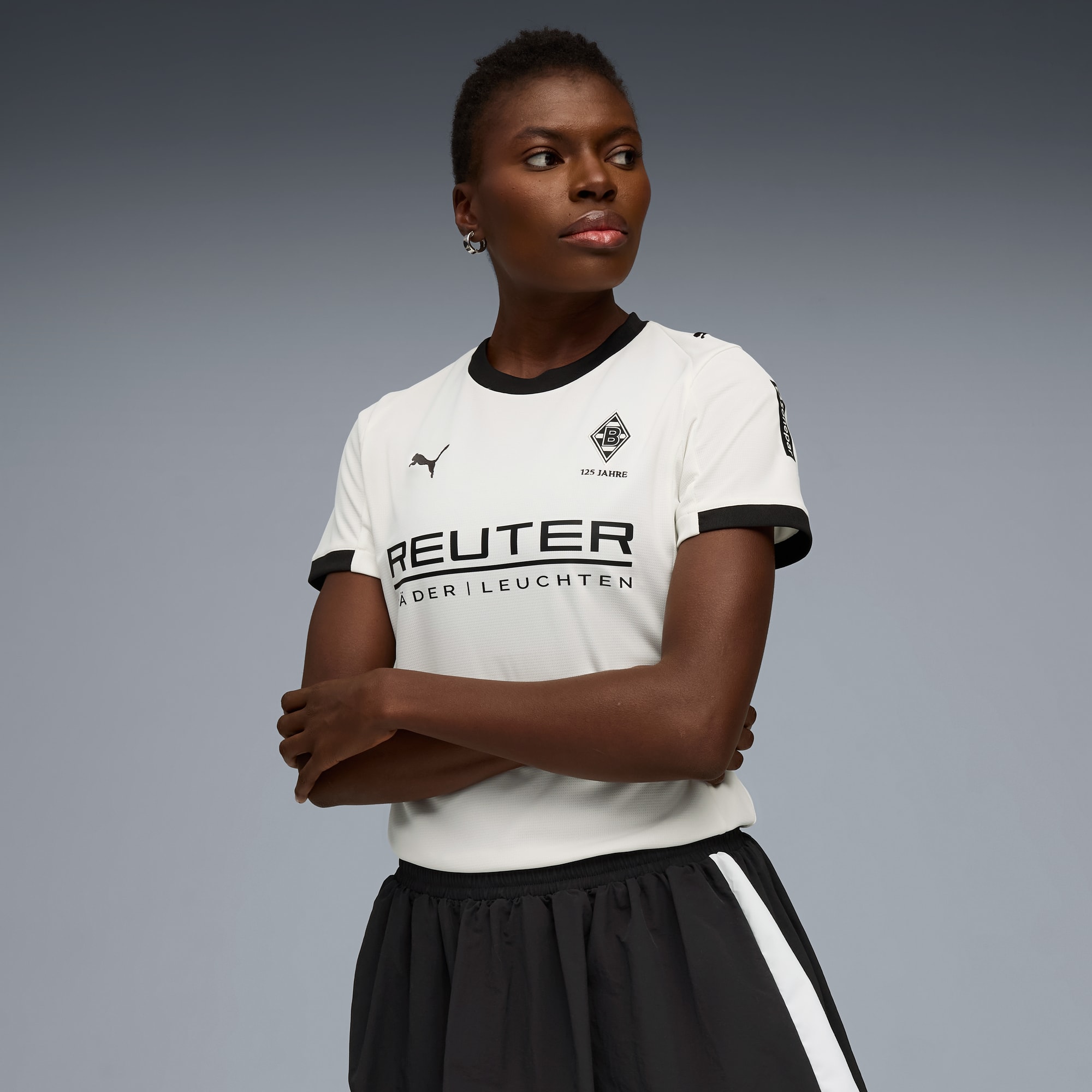 Sur fond gris, un mannequin aux bras crois&eacute;s porte un t-shirt blanc et une jupe noire.