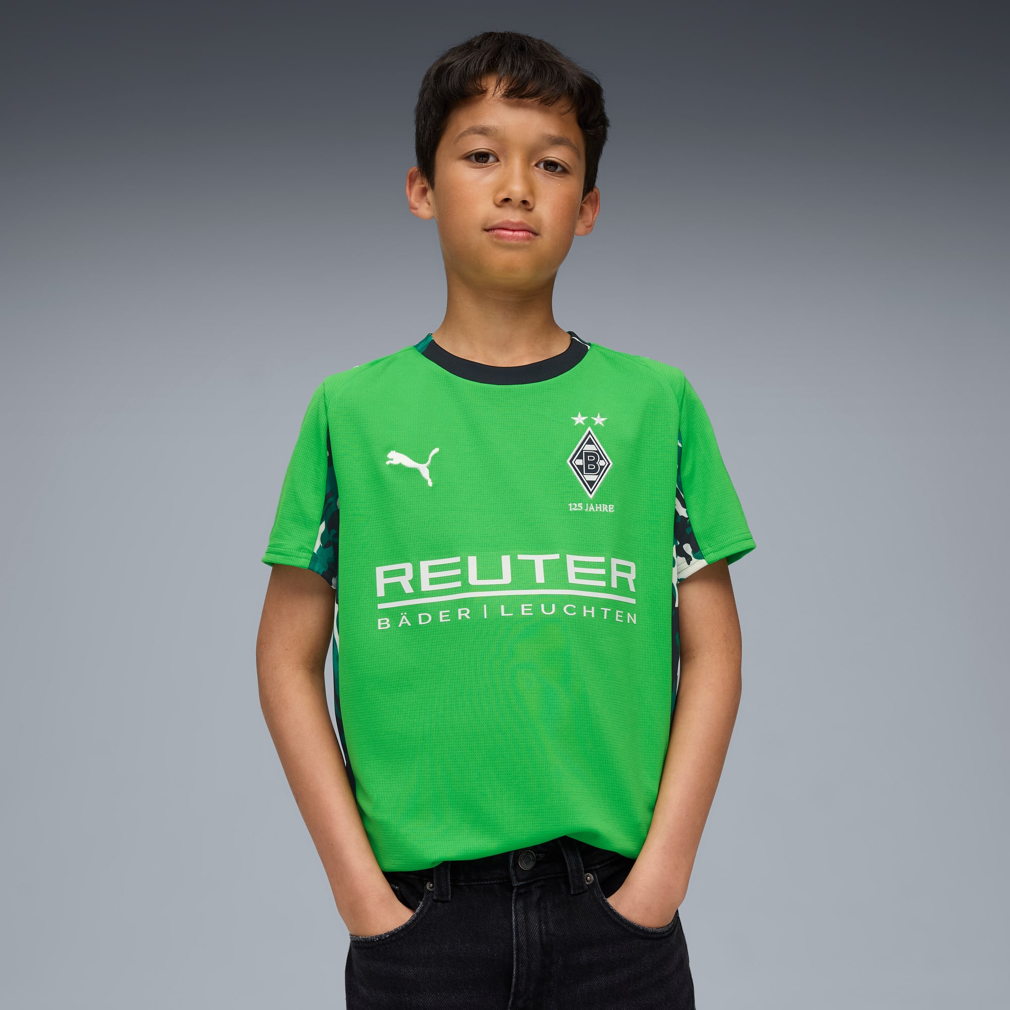Un bambino posa su sfondo grigio, indossando una maglia da calcio verde e jeans scuri.