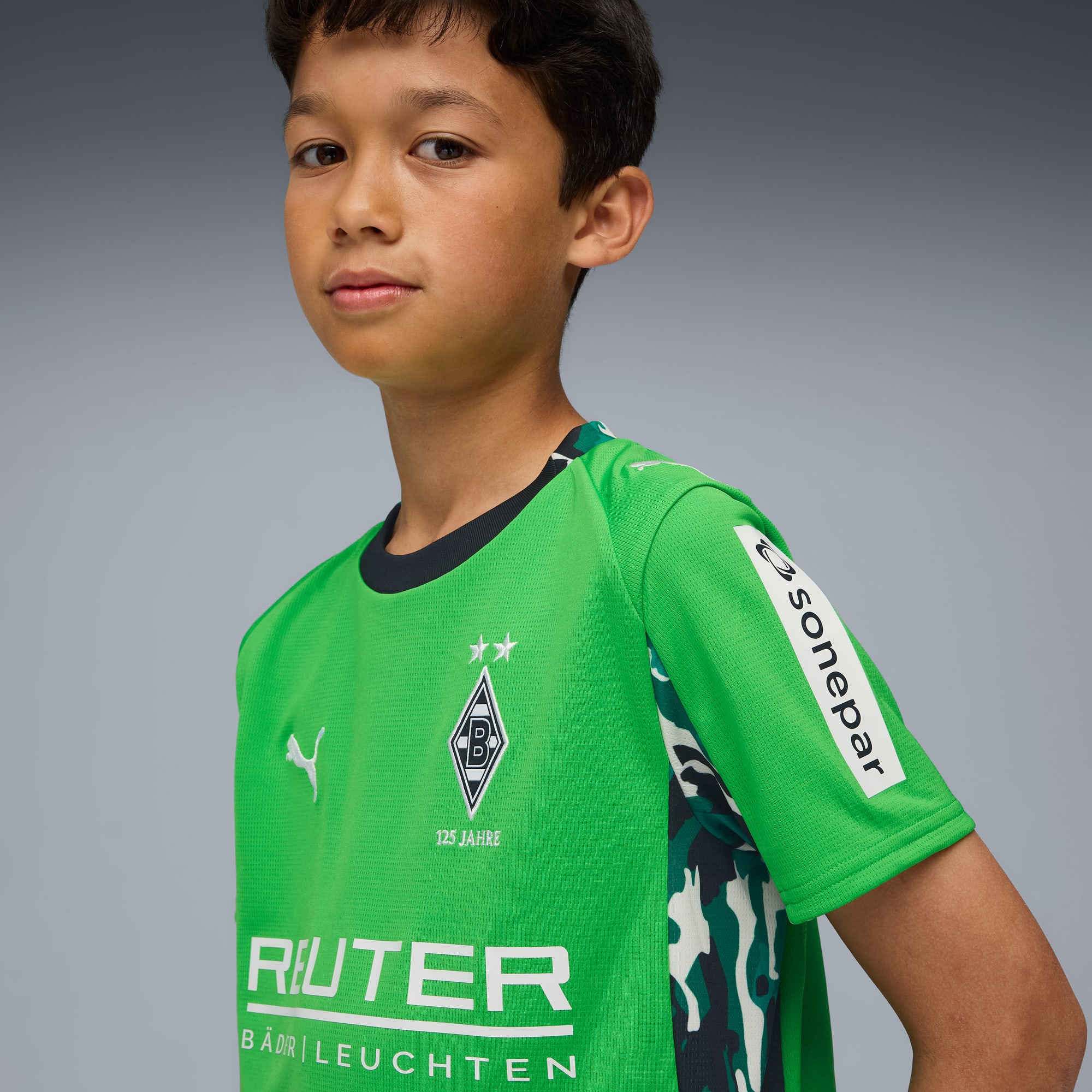 Tegen een grijze achtergrond draagt een kindmodel een groen voetbalshirt met sponsorlogo's.