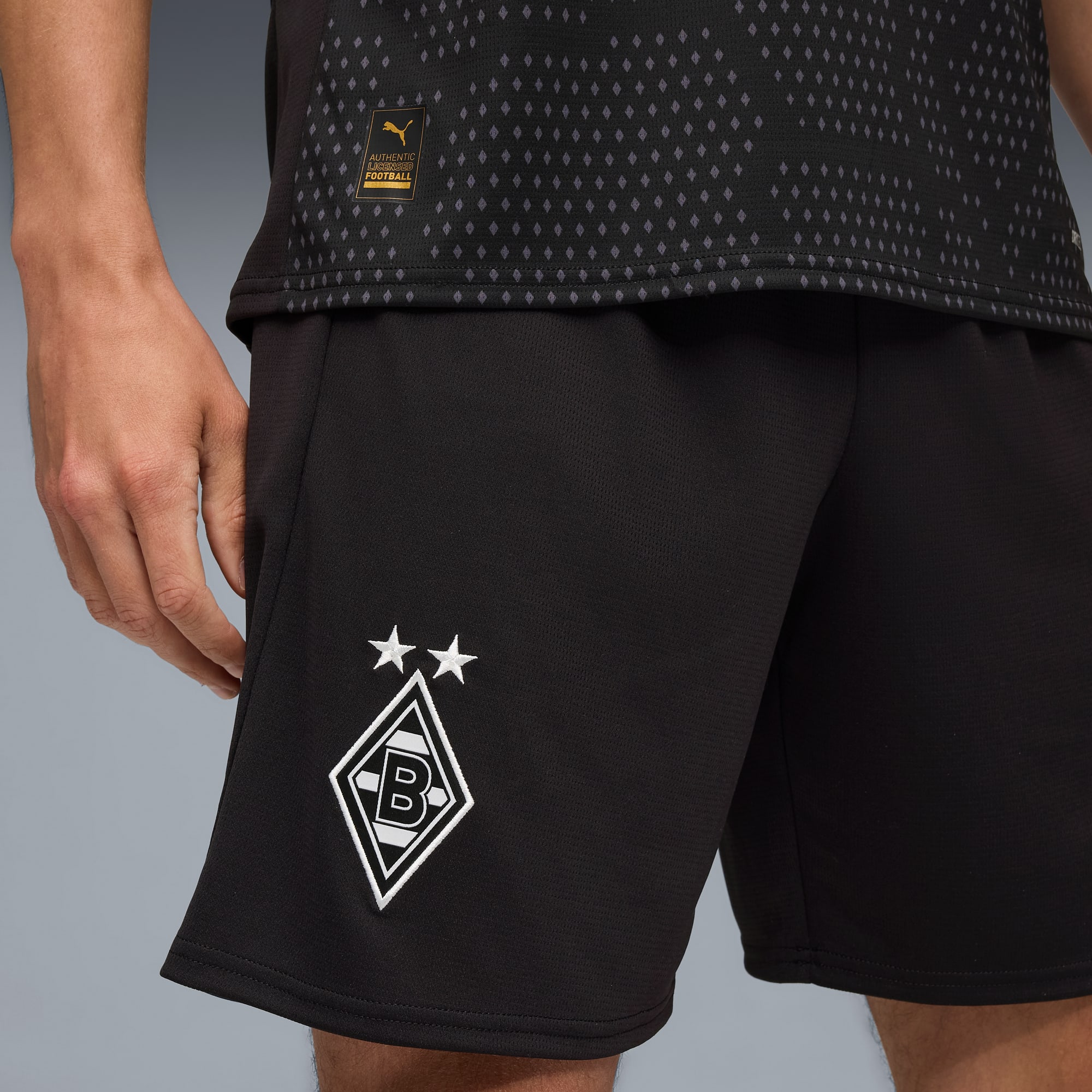 Eine Person trägt ein schwarz-rautenförmiges Oberteil und schwarze Shorts mit weißem Logo vor grauem Hintergrund.