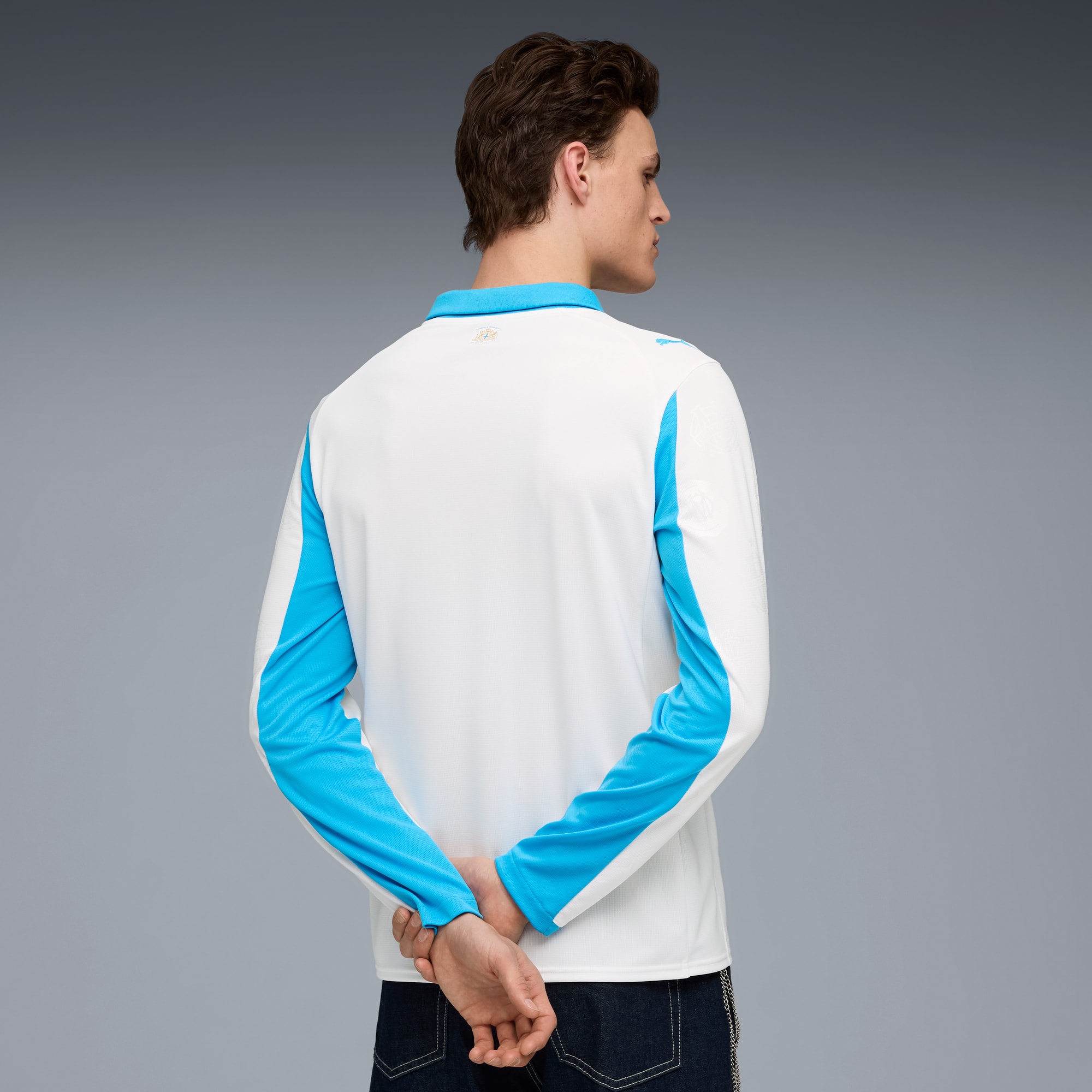 Een model tegen een grijze achtergrond, draagt een wit shirt met lange mouwen en blauwe accenten, handen gekruist.