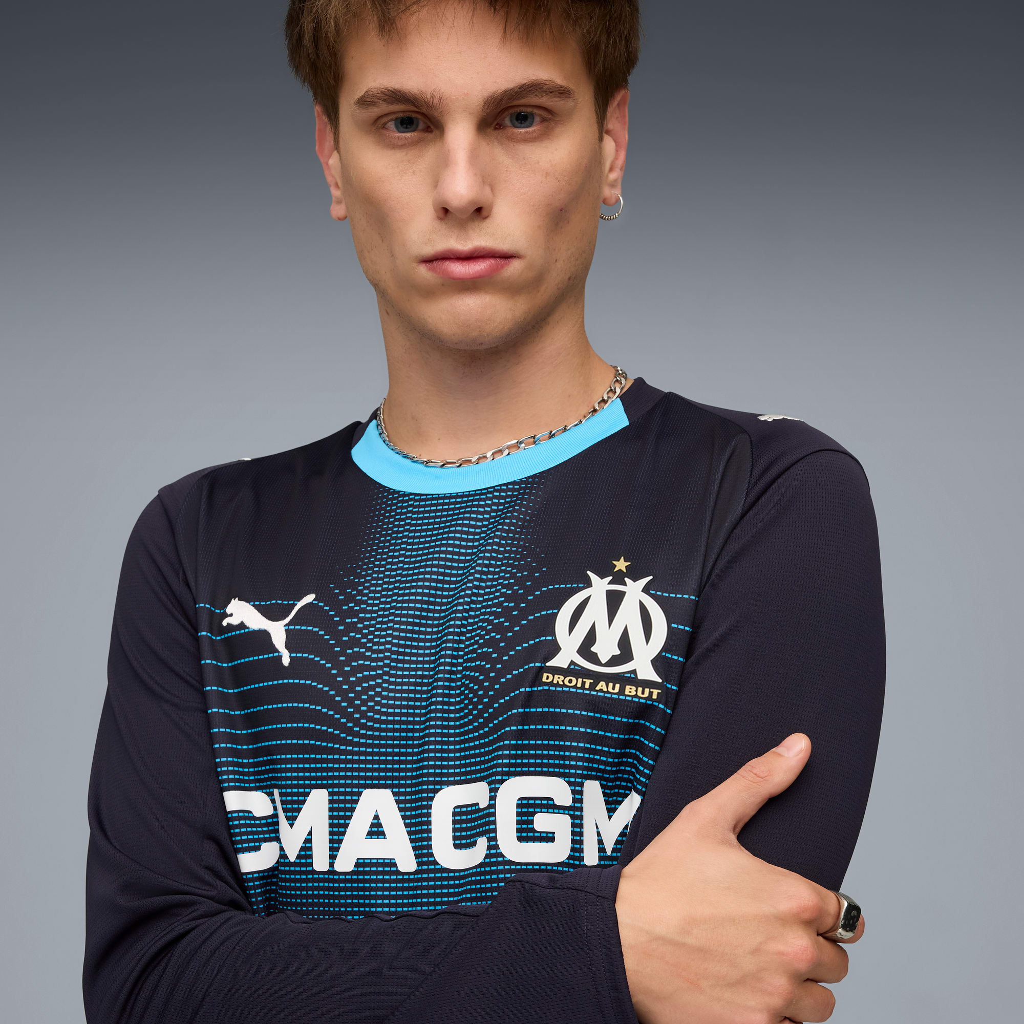 Een model poseert tegen een grijze achtergrond, gekleed in een blauw-zwart shirt met lange mouwen.