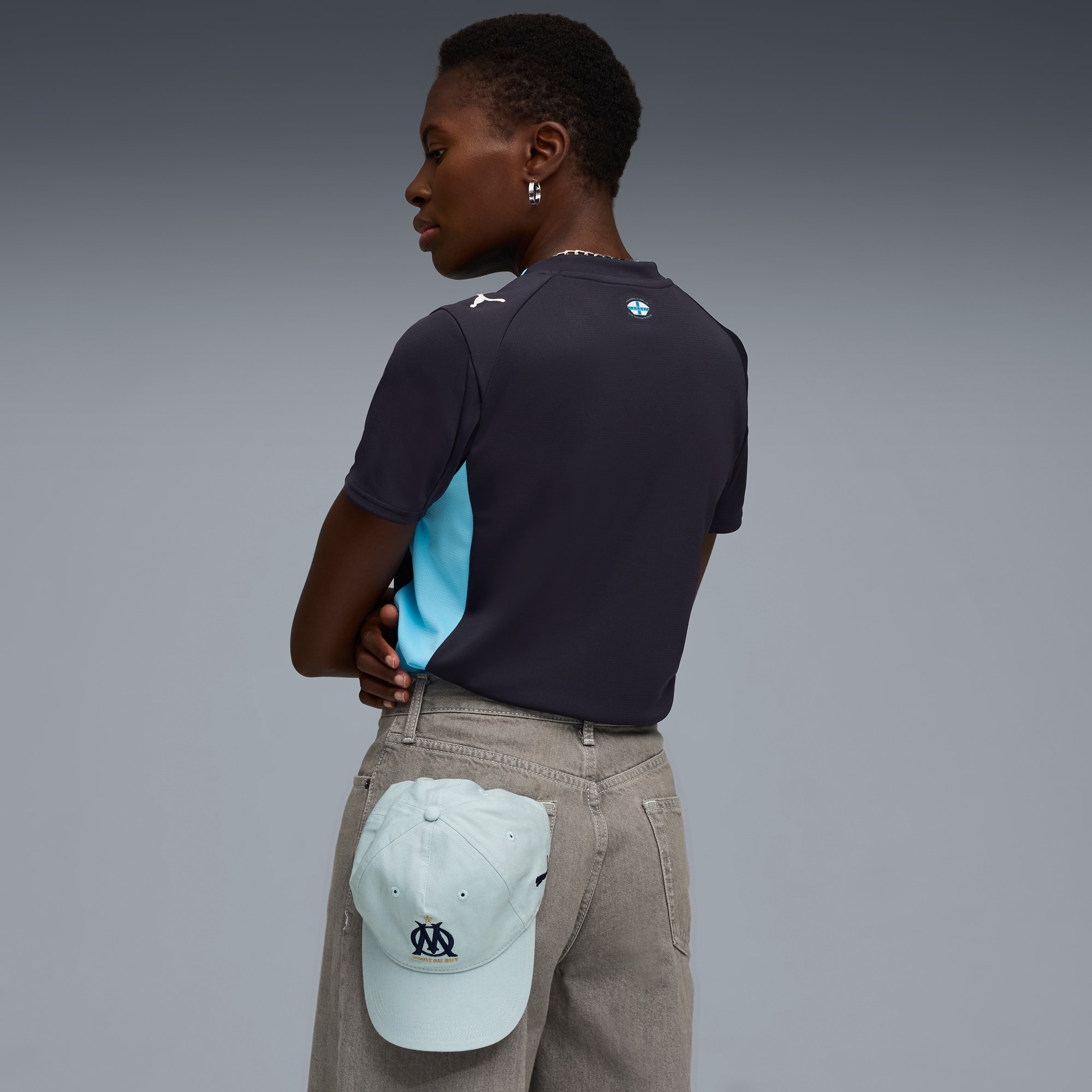 Modella in posa su sfondo grigio con top, pantaloni e cappellino in tasca.