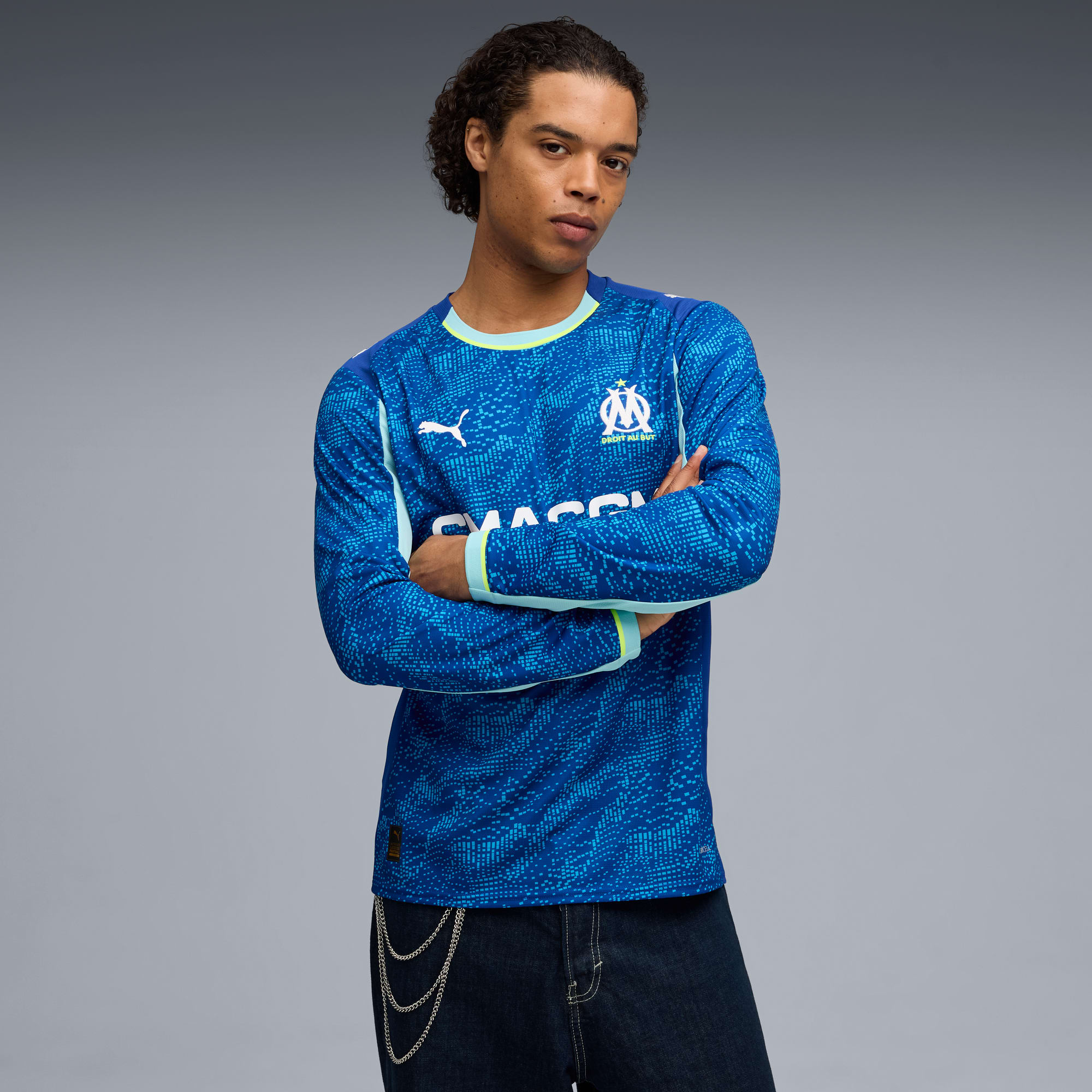 Un mannequin pose, bras croisés, sur un fond gris, vêtu d'un maillot de foot bleu.