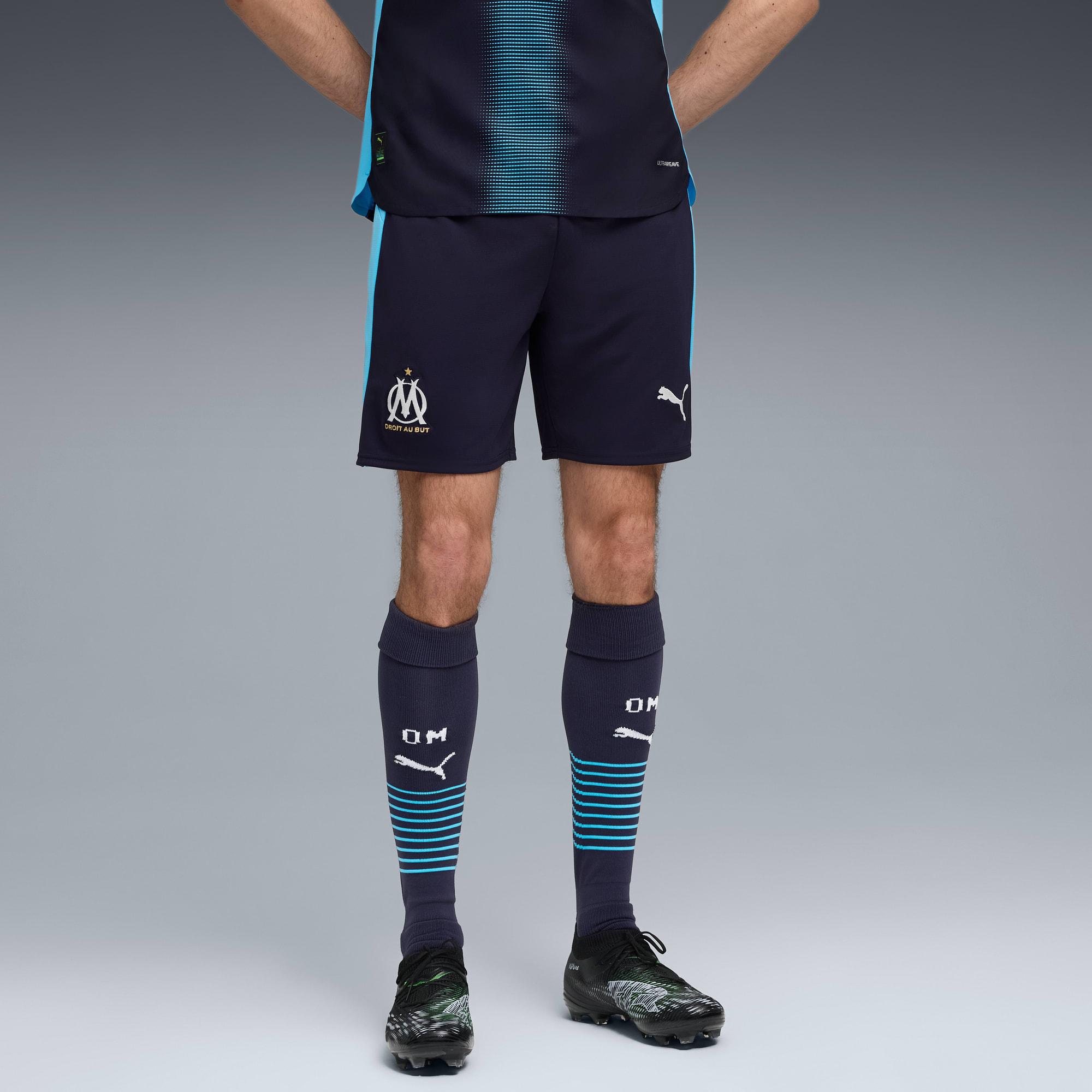 Un modèle pose sur fond gris, portant un kit de football avec short et chaussettes bleu marine.