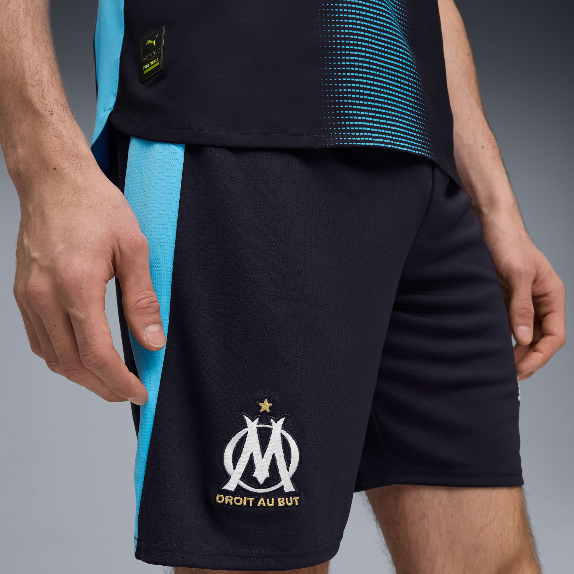 Auf Grau trägt ein Model ein schwarzes Top und Shorts mit hellblauen Akzenten und Teamwappen.