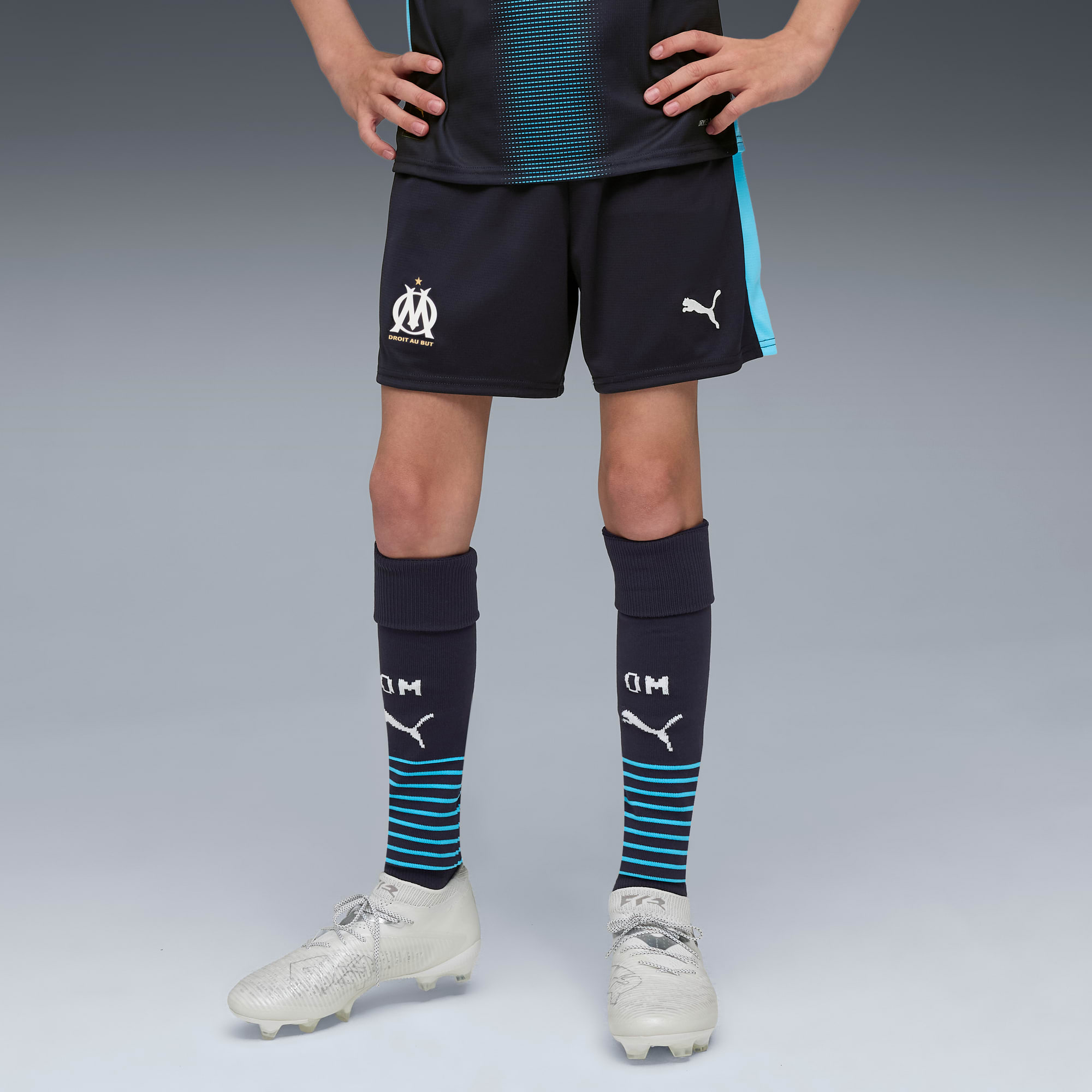 Vor grauem Hintergrund trägt ein Model ein Fußballtrikot mit dunklen Shorts, Socken und weißen Laufschuhen.