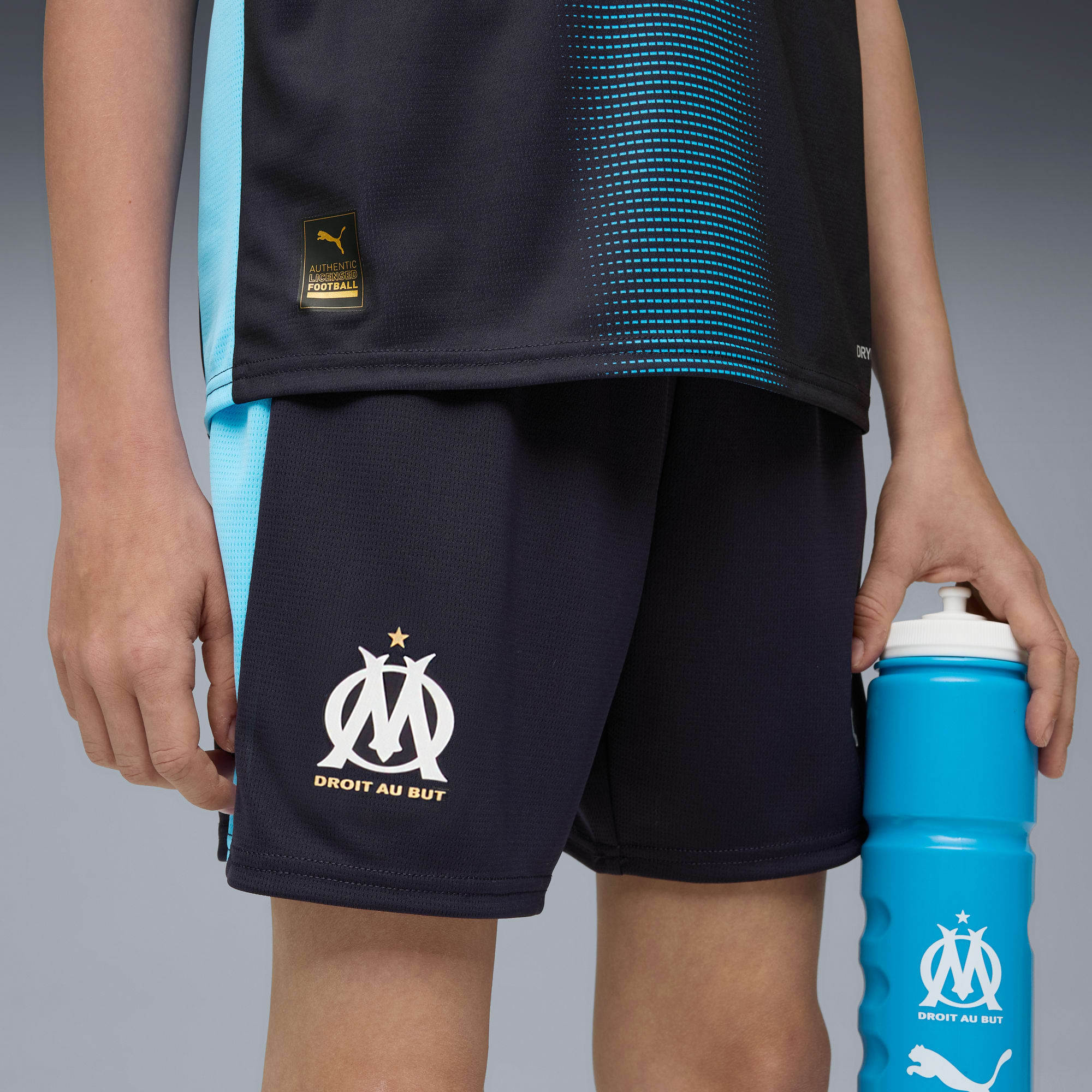 Ein Model in schwarz-blauer Sportbekleidung hält eine blaue Wasserflasche vor grauem Hintergrund.